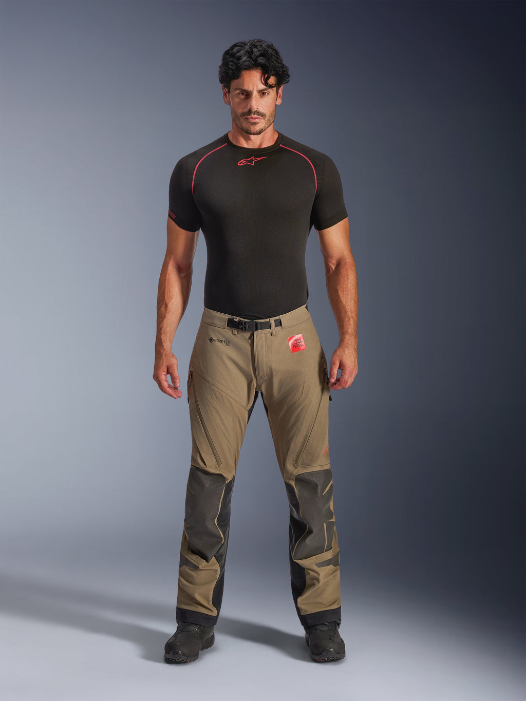 Männliches Model trägt Alpinestars Nazca 3L GORE-TEX PRO Hose in Walnussbraun und Schwarz, technische Adventure-Fahrausrüstung mit Superfabric-Knieverstärkungen und einer magnetischen Gürtelschnalle, kombiniert mit einem schwarzen Alpinestars Base-Layer-Shirt, Studio-Frontansicht.