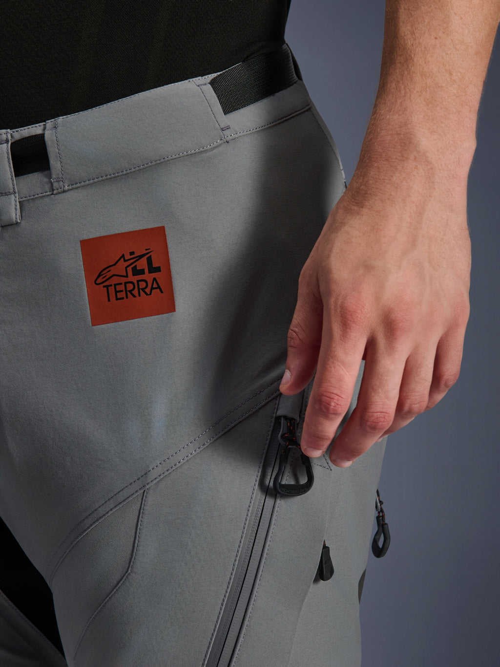 Alpinestars Nazca 3L GORE-TEX PRO Hose, Adventure-Motorradhose, Dunkelgrau, Nahaufnahme des oberen Oberschenkels mit rotbraunem All Terra Logo-Aufnäher, Cargo-Tasche mit Reißverschluss und schwarzen Zippern, strapazierfähige Textilkonstruktion für All-Terrain-Fahrten