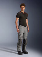 Männliches Model in der Alpinestars Nazca 3L GORE-TEX PRO Hose, dunkelgraue und schwarze Adventure-Motorradhose, mit verstärkten Knieeinsätzen und Belüftungsreißverschlüssen, getragen mit einem schwarzen Base-Layer-Oberteil, technische Ausrüstung für extreme All-Terrain-Fahrten.
