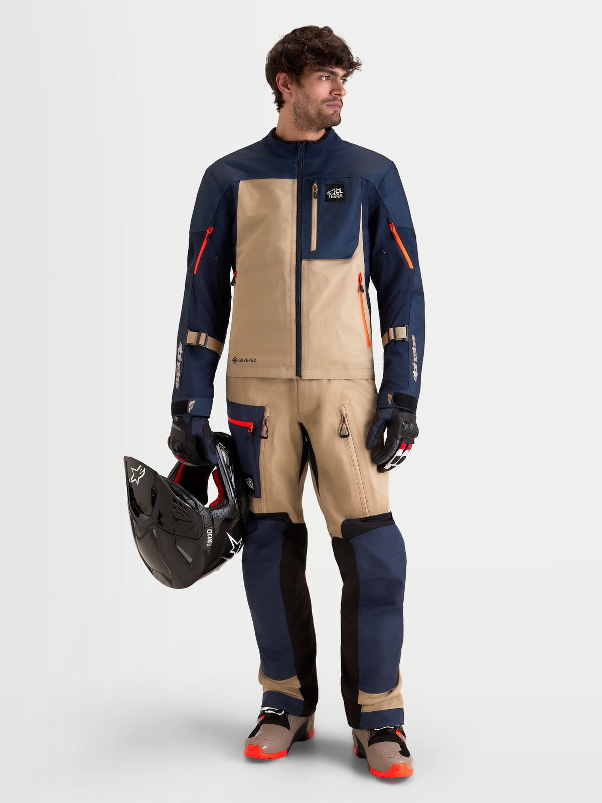 Alpinestars Arenis Gore-Tex Hose, Herren-Adventure-Motorradhose in Nachtblau und Aluminium, getragen von einem Model mit passender ADV-Ausrüstung und Helm in der Hand
