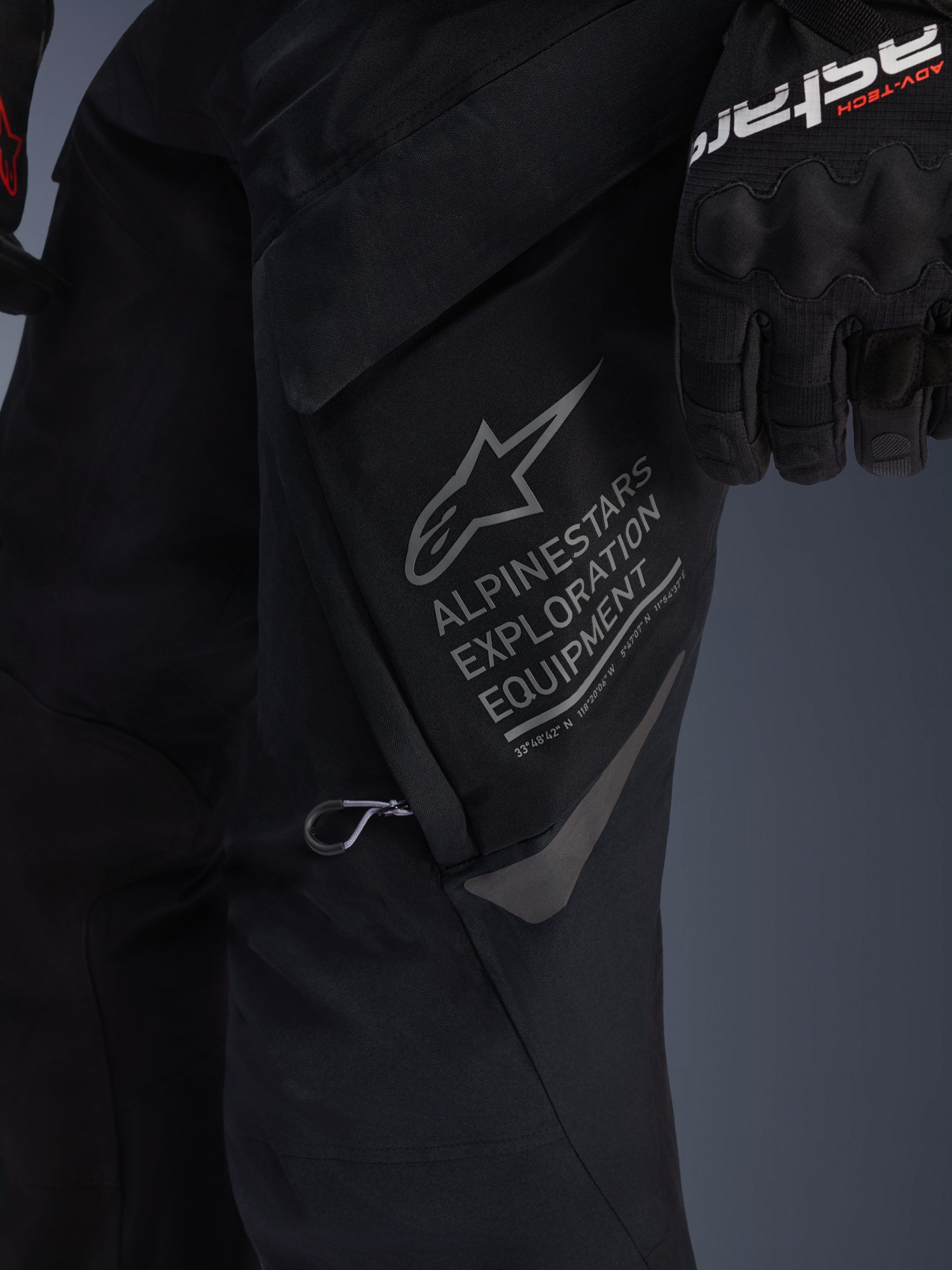 Nahaufnahme der Alpinestars ST-7 2L GORE-TEX Hose in Schwarz, Detailansicht der großen Cargo-Tasche am Oberschenkel mit grauem Alpinestars Exploration Equipment Branding, Stern-Logo und geografischen Koordinaten, Hochleistungs-Textilkonstruktion für Adventure-Touring.