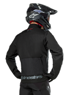 Back view of a rider wearing the Alpinestars Pro-Dura Jacket in Black, durable ripstop textile enduro jacket, featuring a large rear cargo pocket with gray Alpinestars branding across the lower back, paired with a black motocross helmet.