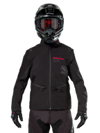 Motocross rider wearing Alpinestars Lite-Dura Softshell Jacket in Black, featuring red and grey logo details on the chest, black helmet with red-tinted goggles, black gloves and pants, front view of technical off-road riding apparel