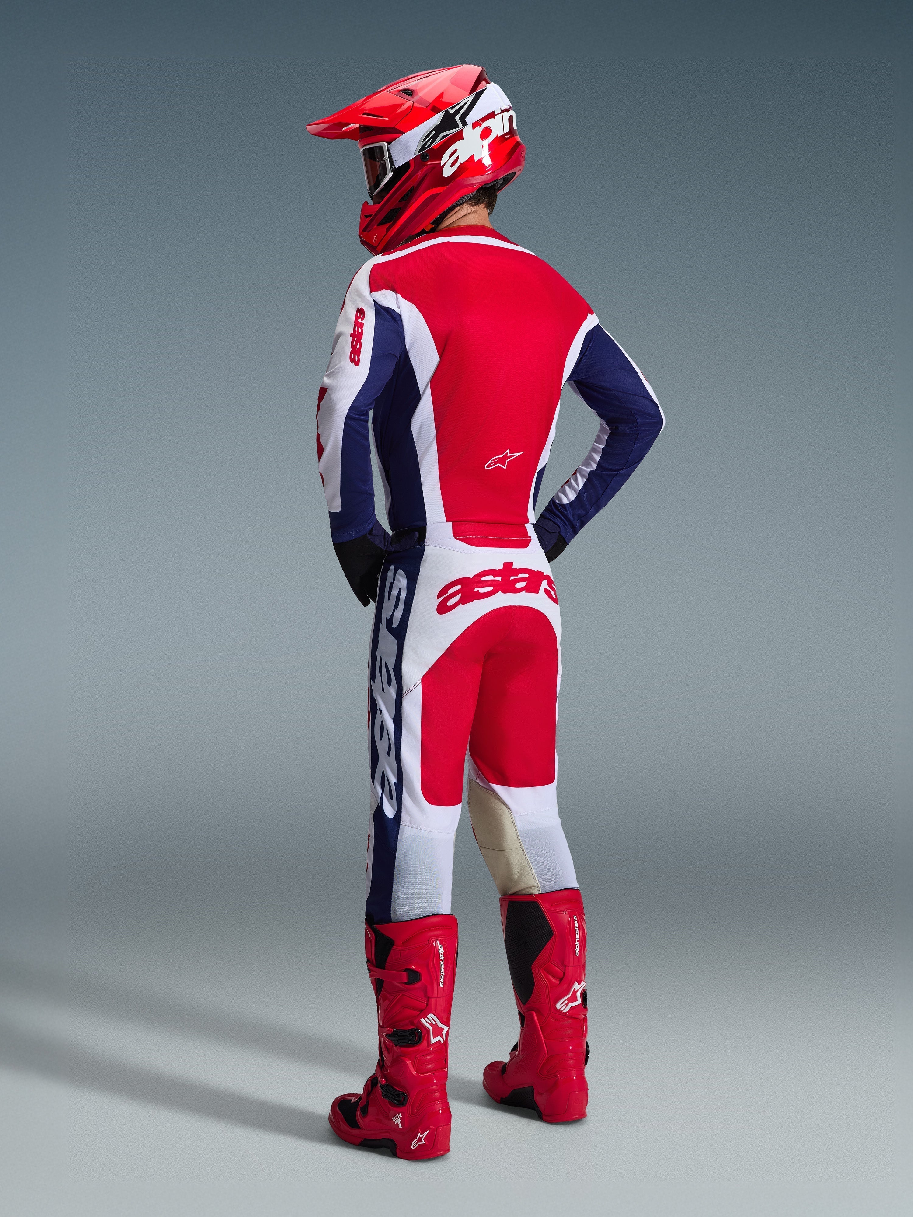 Motocross-Fahrer in der Rückansicht, Alpinestars 2026 Racer Riway Hose in Weiß, Blau und Rot, kombiniert mit passendem Trikot, rotem Helm und leuchtend roten Stiefeln, markantes Branding auf der Sitzfläche, sportliche Offroad-Passform