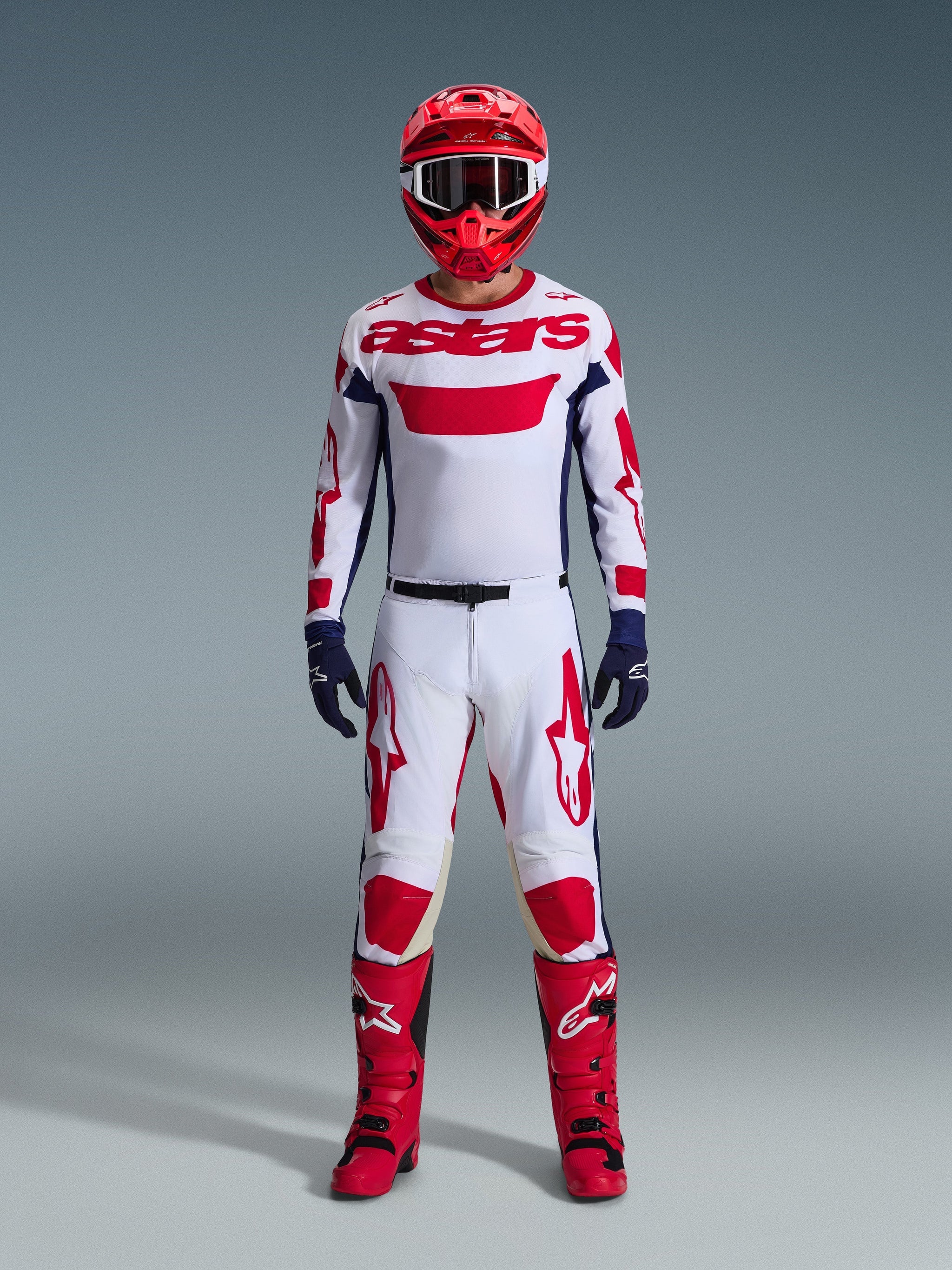 Motocross-Fahrer in kompletter Rennausrüstung, weißes Trikot mit rotem Alpinestars-Branding und blauen Akzenten, Alpinestars 2026 Racer Riway Hose in Weiß-Blau-Rot, roter Helm mit passender Brille, rote und schwarze Stiefel, marineblaue Handschuhe, abgestimmtes Outfit für den Offroad-Rennsport