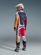 Motocross-Fahrer in der Rückansicht in kompletter Rennausrüstung, Alpinestars 2026 Racer Portl Hose in Rot, Hellgrau und Dunkelgrau, passendes Trikot mit orangefarbenen und weinroten Ärmeln, mehrfarbiger Grafikhelm, graue und schwarze Offroad-Stiefel mit pinkfarbenen Akzenten, professionelles Motocross-Outfit