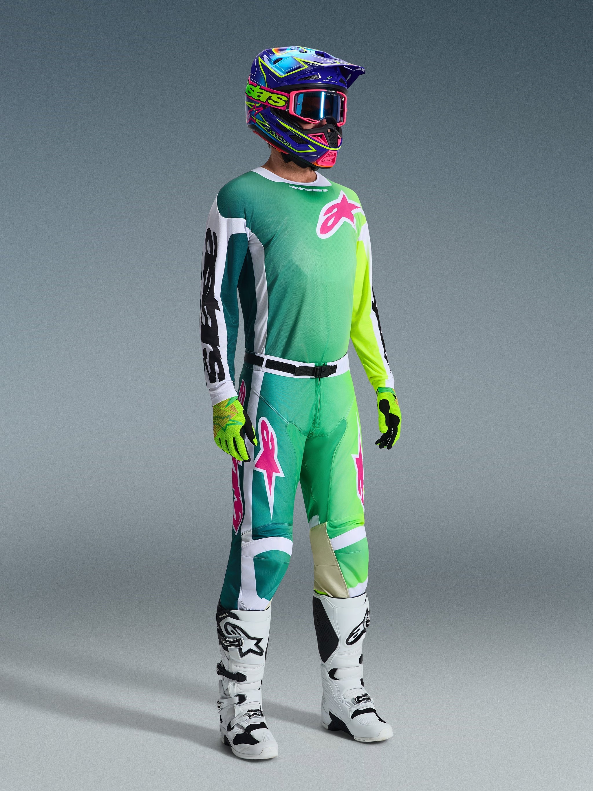 Motocross-Fahrer mit Alpinestars 2026 Racer Portl Hose und passendem Trikot in Grün, Weiß und Schwarz mit pinkfarbenen Logo-Akzenten, bunter Helm, weiß-schwarze Rennstiefel und neongrüne Handschuhe, komplette Ausrüstung für den Offroad-Rennsport