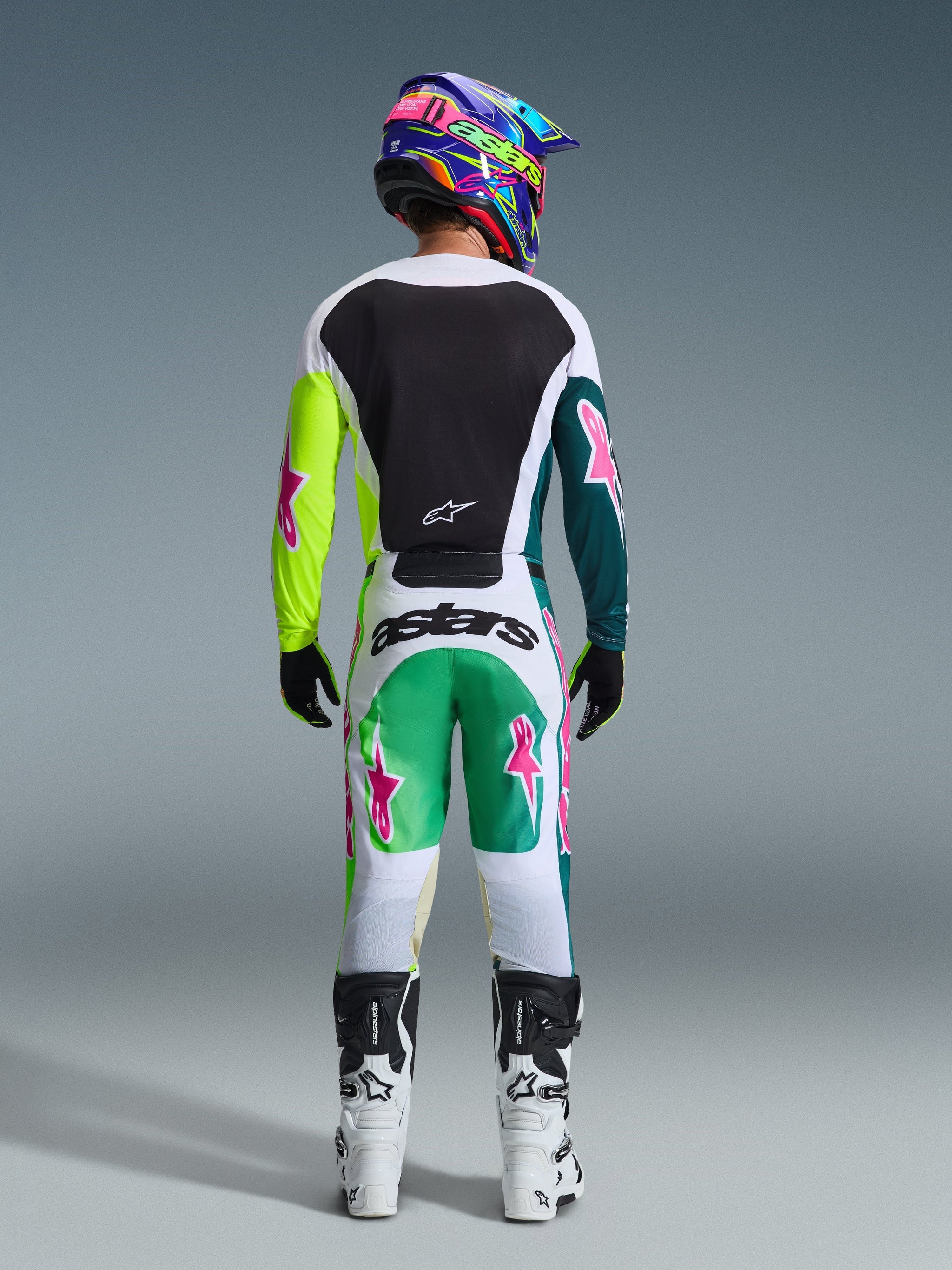Motocross-Fahrer von hinten in der Alpinestars 2026 Racer Portl Hose in Grün-Weiß-Schwarz, passendes Trikot mit pinkfarbenen Stern-Logos, mehrfarbiger „astars“-Helm und weiß-schwarze Stiefel, abgestimmte Offroad-Rennausrüstung.