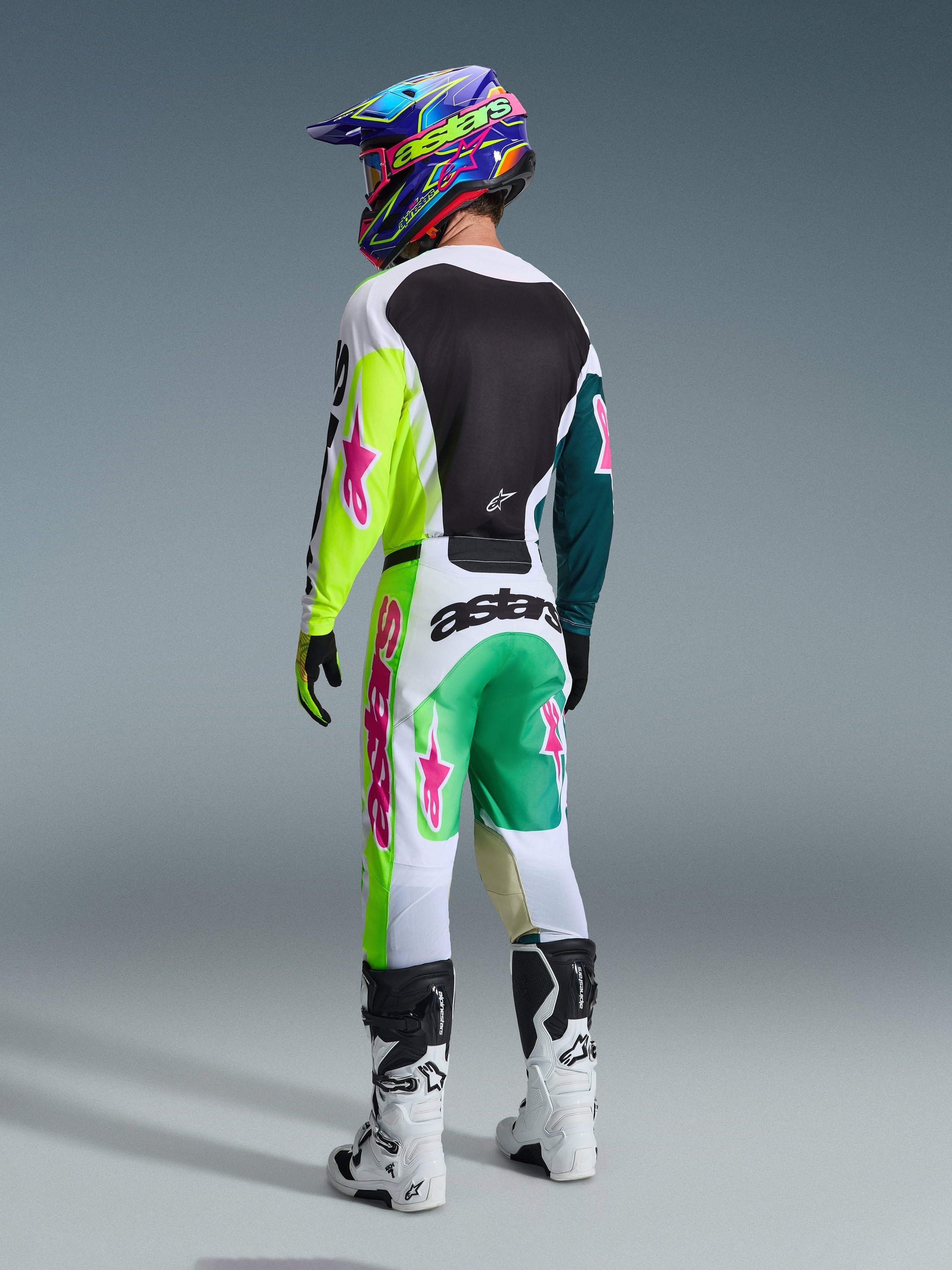 Motocross-Fahrer von hinten in Alpinestars 2026 Racer Portl Hose in Grün Weiß Schwarz, passendes Trikot mit limonengrünen und pinken Akzenten, bunter Helm, weiße und schwarze Stiefel, komplette Offroad-Rennausrüstung.