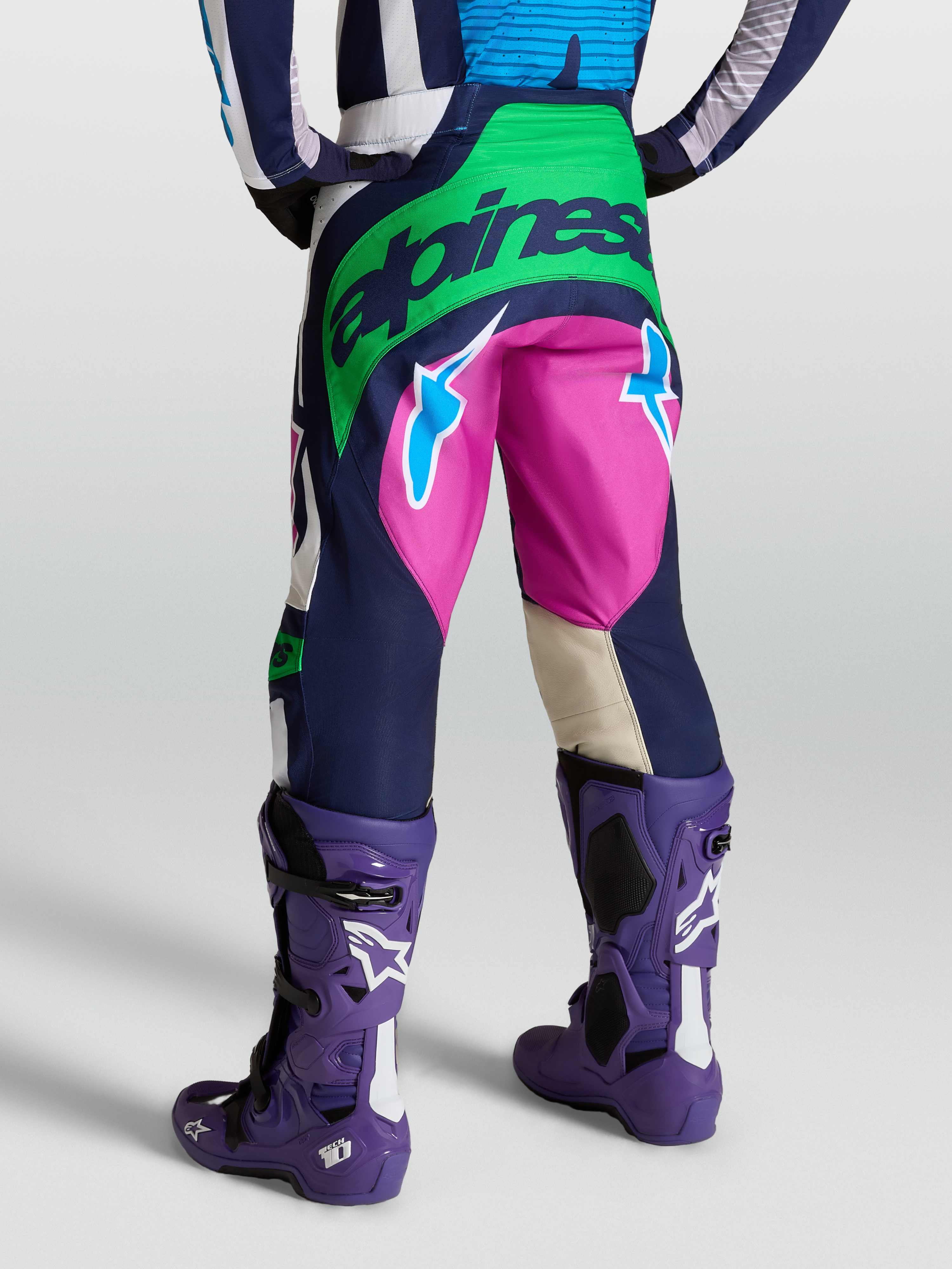 Alpinestars Techstar Maven Hose, Motocross-Ausrüstung in Blau, Grün und Pink, Rückansicht am Model mit violetten Tech 10 Stiefeln, strapazierfähige Offroad-Bekleidung.