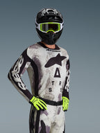 Alpinestars 2026 Racer Lahnd Trikot, Motocross-Trikot, Sand Camo mit grauem und schwarzem Muster, großes Astars-Branding auf der Brust, getragen mit schwarzem Helm, neongelber Brille und Handschuhen, Performance-Offroad-Rennausrüstung