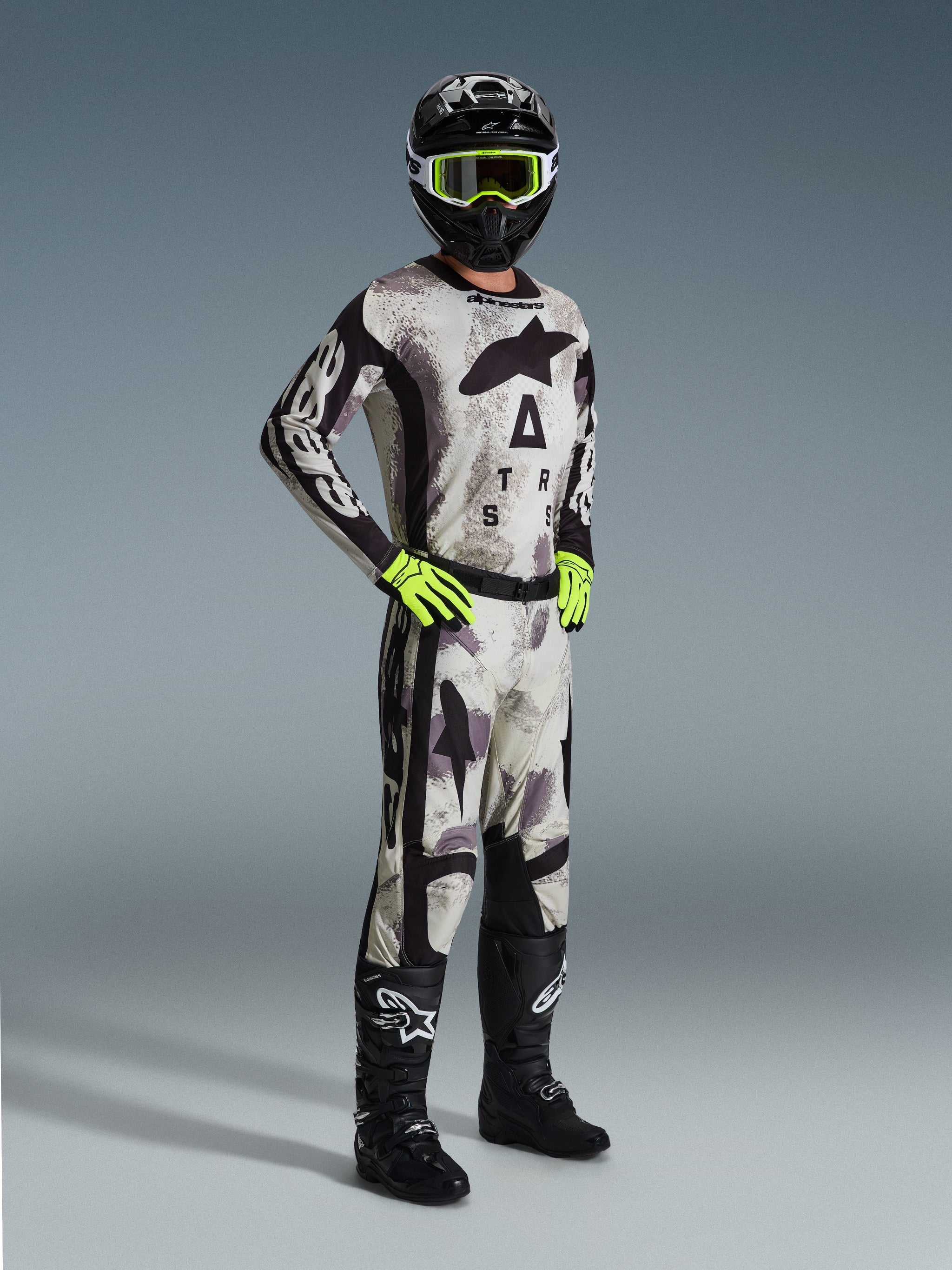 Motocross-Fahrer in kompletter Alpinestars Rennausrüstung, 2026 Racer Lahnd Hose in Sand Camo, passendes Camo-Trikot mit auffälligem Branding, schwarzer Helm mit neongelber Brille, neongrüne Handschuhe und schwarze Stiefel, koordiniertes Profi-Outfit für Offroad-Performance.