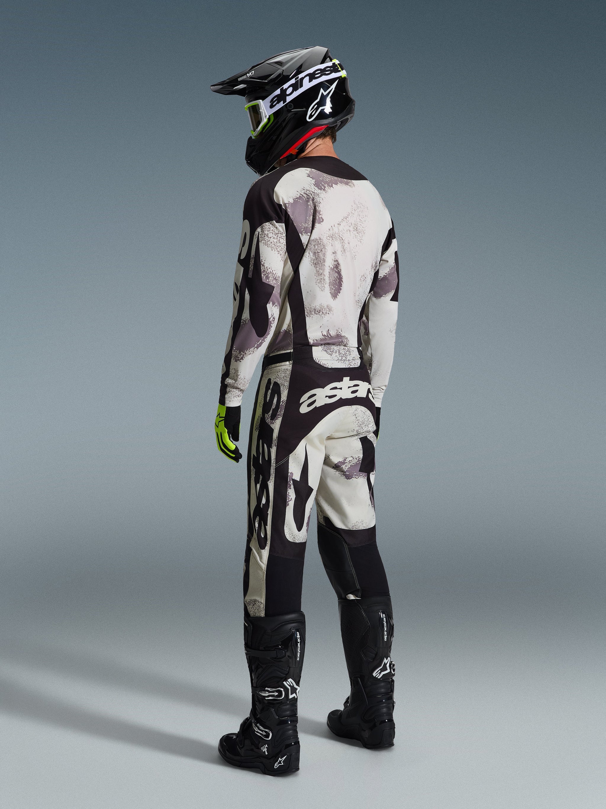 Motocross-Fahrer trägt die Alpinestars 2026 Racer Lahnd Hose in Sand Camo und Grau, komplettes Gear-Set mit passendem Jersey, schwarzem Helm und schwarzen Stiefeln, Rückansicht, die das markante Branding und das ergonomische Design hervorhebt.