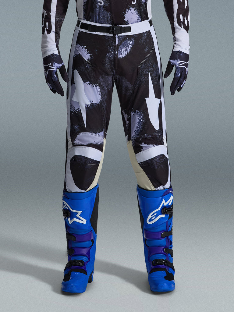Alpinestars 2026 Racer Lahnd Hose, Motocross-Hose, Iron Camo, getragen mit passenden Camouflage-Handschuhen und leuchtend blauen und lila Stiefeln, mit stilisierten weißen Stern-Grafiken auf einem dunklen Camo-Muster, technischer Magnetverschluss, professionelle Offroad-Rennausrüstung.