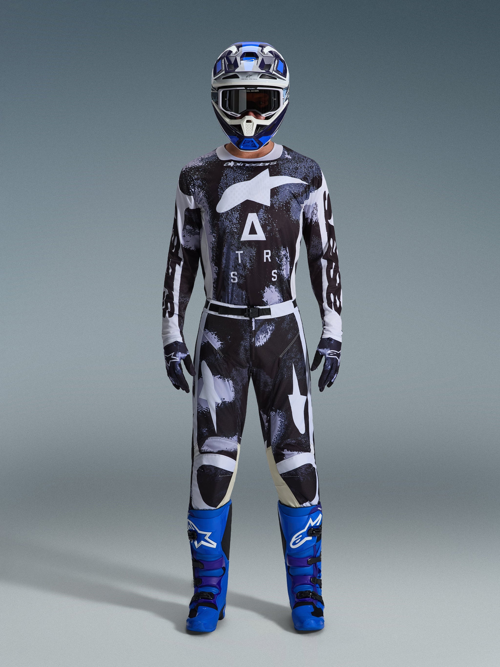 Motocross rider wearing Alpinestars 2026 Racer Lahnd Pants in Iron Camo, gray and black camouflage design, matching jersey, blue and white helmet with goggles, and bright blue boots, coordinated motocross gear set.