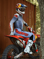 Motocross rider sitting on a red dirt bike, wearing the Alpinestars 2025 Supertech Lipan Gear Set in Gray and Orange Fluo, light blue pants with bold orange Astars logo, matching jersey, white motocross boots, and a Red Bull helmet, coordinated professional racing gear