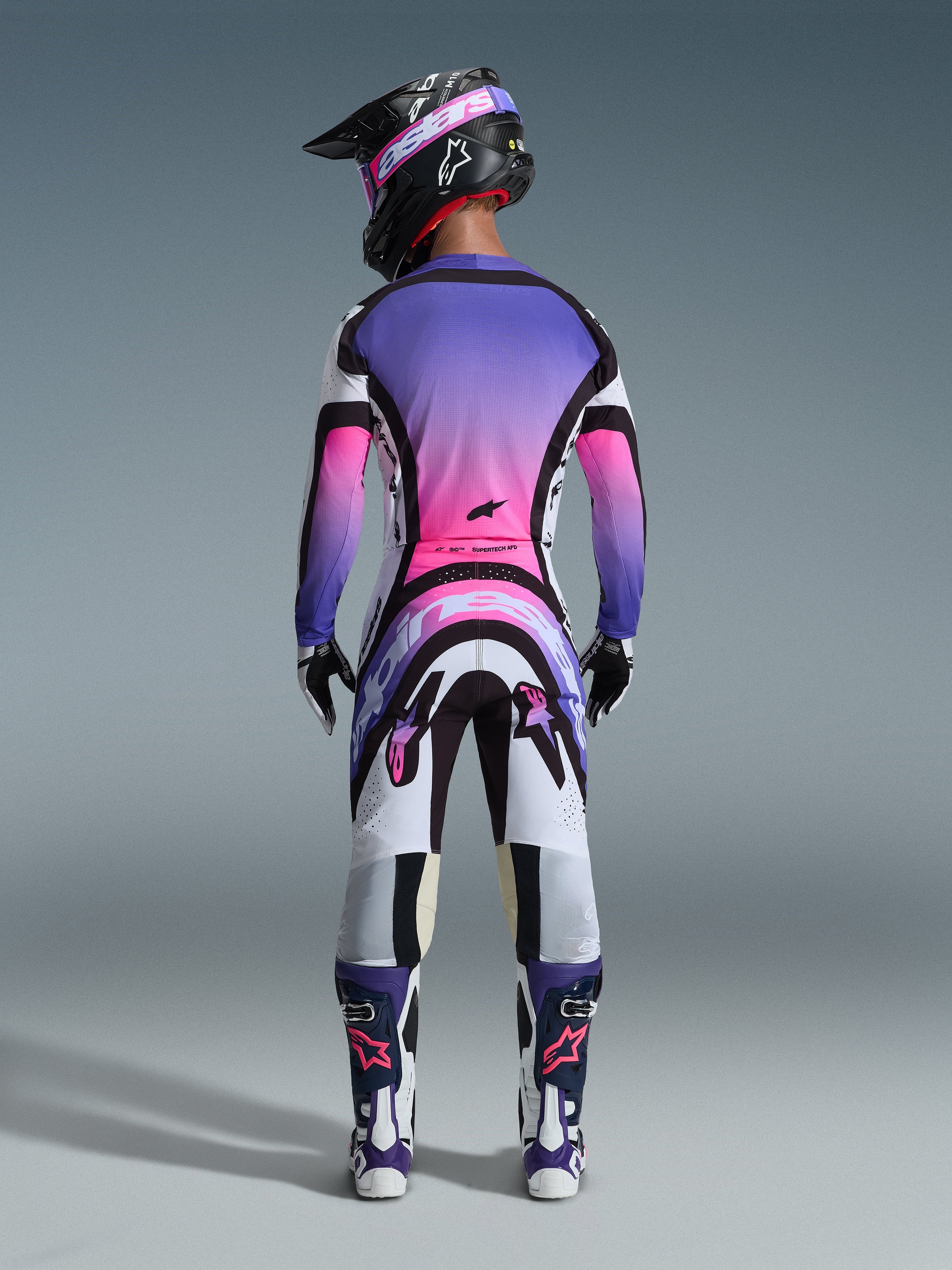 Motocross-Fahrer in der Rückansicht, komplette Rennausrüstung, Trikot mit lila-pinkem Farbverlauf, Alpinestars 2026 Supertech AFD Vista Hose in Weiß, Lila und Schwarz, schwarzer Helm mit lila Brille, weiß-lila Stiefel, farblich abgestimmtes Offroad-Rennset.