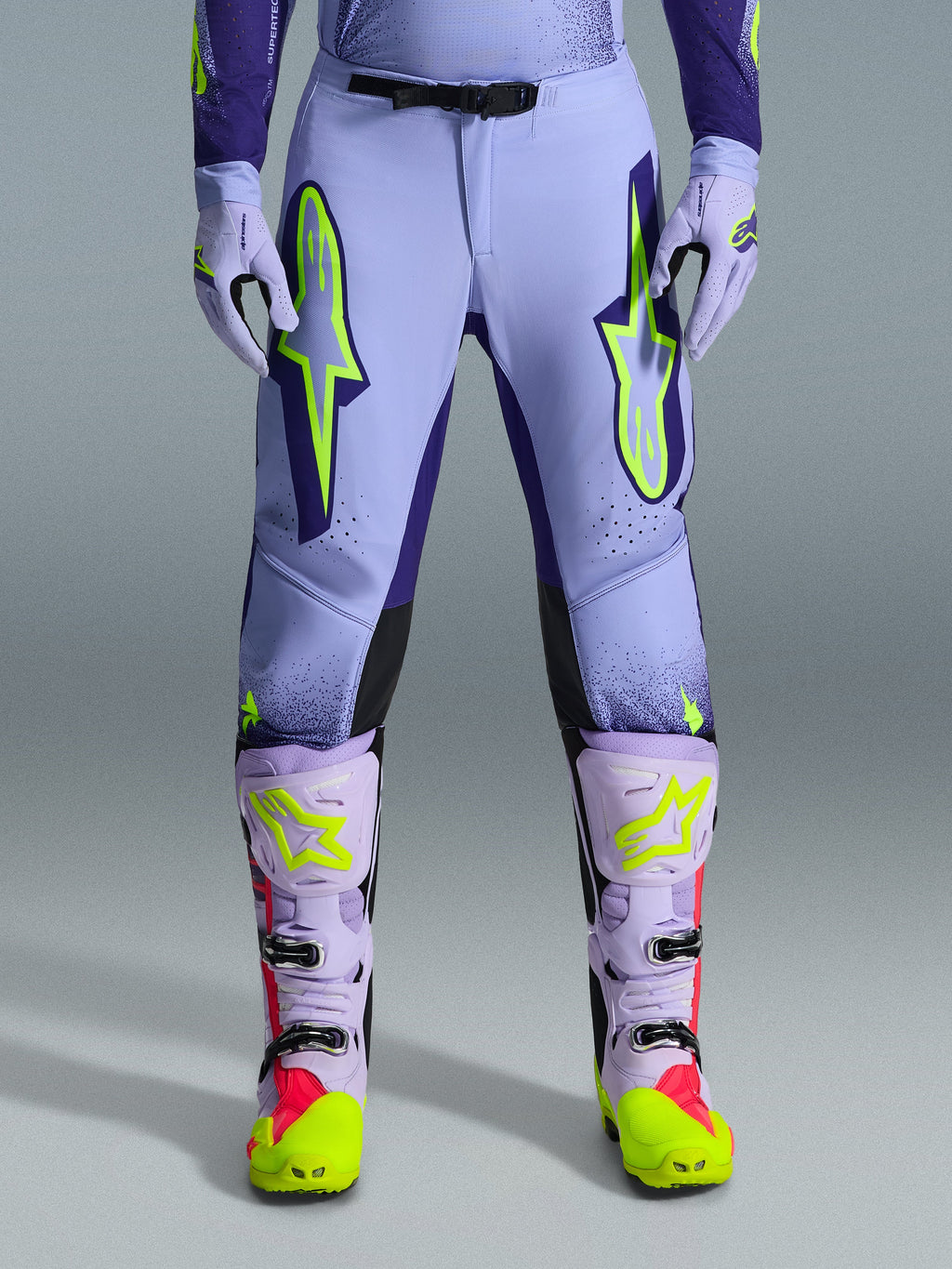 Alpinestars 2026 Supertech Scenz Hose, Motocross-Hose, Lila Gelb Fluo, mit großen neongelben Logos auf den Oberschenkeln, getragen mit passenden lila-gelben Motocross-Stiefeln und -Handschuhen, athletisches Performance-Design für den Offroad-Rennsport