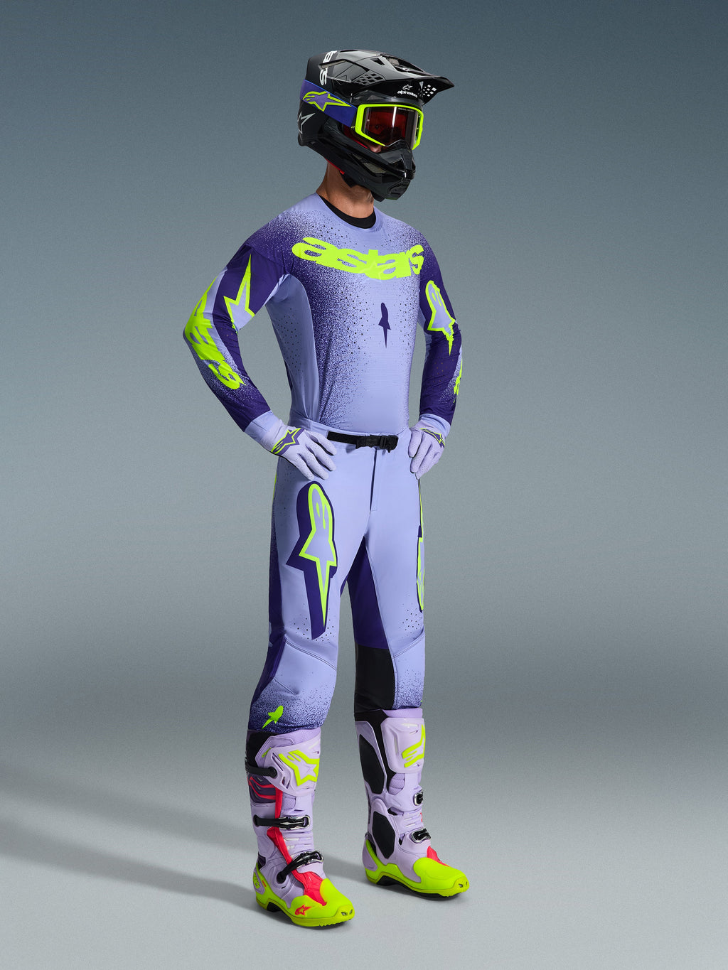 Alpinestars 2026 Supertech Scenz Hose, Motocross-Hose, Lila und Gelb Fluo, getragen von einem Fahrer in einem kompletten Rennoutfit mit passendem Jersey mit Farbverlauf, fliederfarbenen Handschuhen und abgestimmten gelben und lila Stiefeln, inklusive schwarzem Helm und Schutzbrille.