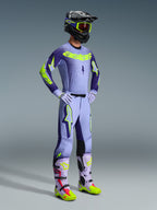 Alpinestars 2026 Supertech Scenz Hose, Motocross-Hose, Lila und Gelb Fluo, getragen von einem Fahrer in einem kompletten Rennoutfit mit passendem Jersey mit Farbverlauf, fliederfarbenen Handschuhen und abgestimmten gelben und lila Stiefeln, inklusive schwarzem Helm und Schutzbrille.