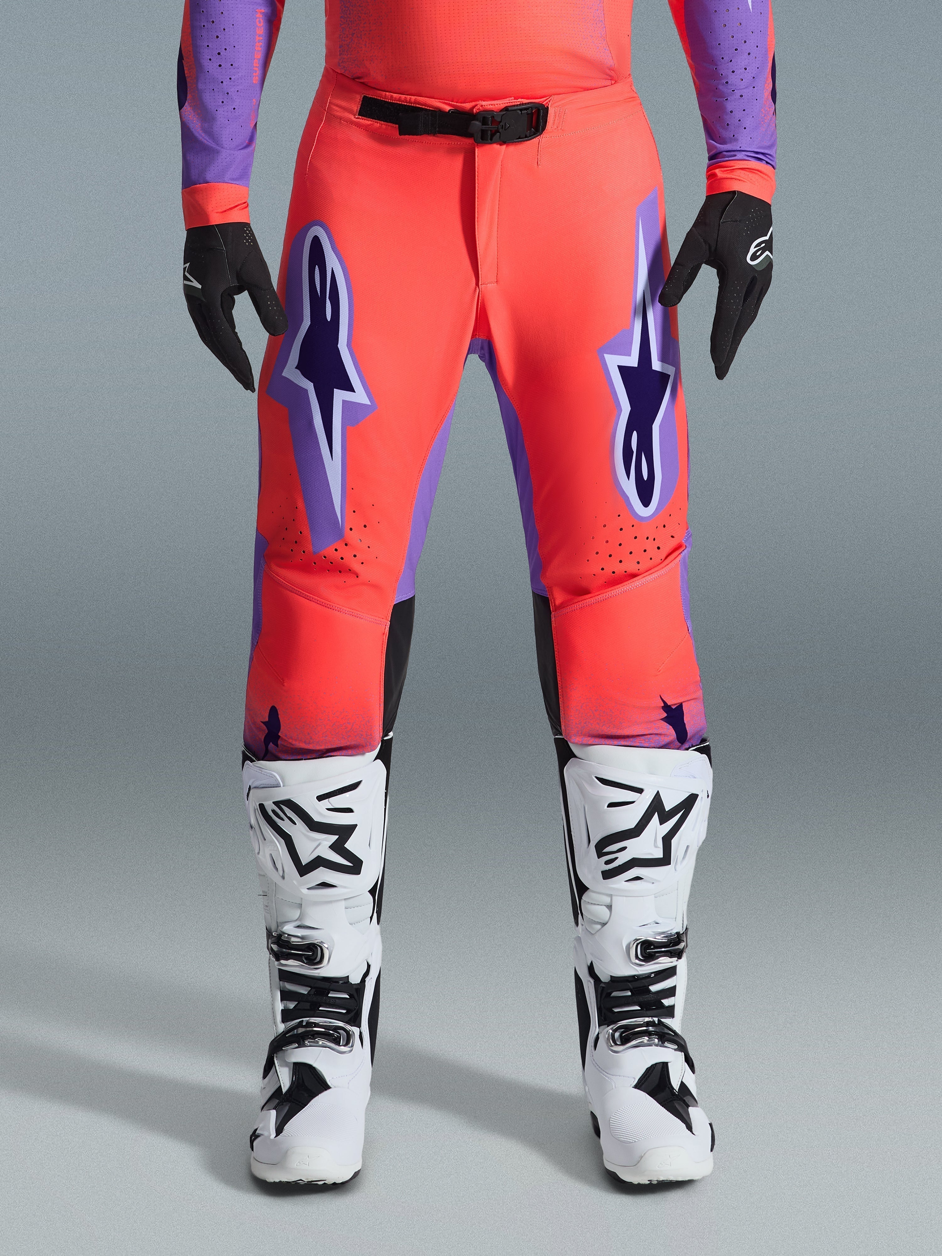 Alpinestars 2026 Supertech Scenz Hose, Motocross-Hose, Hot Coral und Lila, getragen mit passendem Trikot, weißen und schwarzen Stiefeln sowie schwarzen Handschuhen, mit großen lila Astars-Logos auf den Oberschenkeln und einer schwarzen magnetischen Taillenschnalle, professionelle athletische Passform für den Rennsport