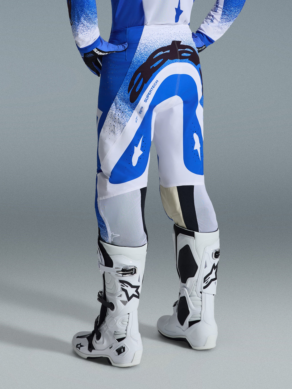 Alpinestars 2026 Supertech Scenz Hose, Motocross-Hose, Ucla-Blau und Weiß, Rückansicht mit großem schwarzem Astars-Logo auf dem Gesäß und „SUPERTECH“-Branding, getragen mit weißen Tech 10 Stiefeln und einem passenden blau-weißen Trikot.