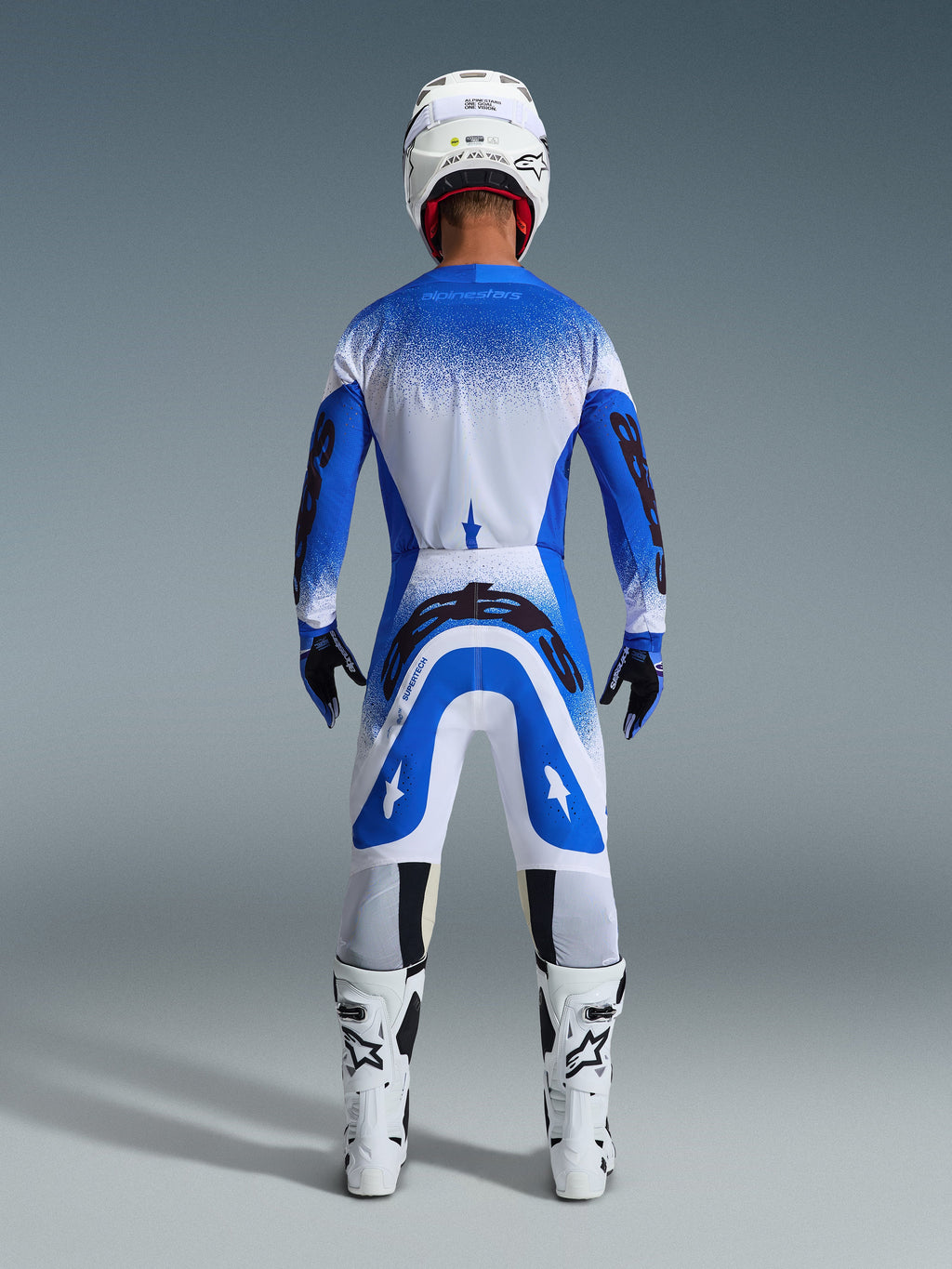 Motocross-Fahrer von hinten in kompletter Rennausrüstung, Alpinestars 2026 Supertech Scenz Hose in Ucla-Blau und Weiß, passendes blau-weißes Trikot mit gesprenkeltem Muster, weißer Helm, weiß-schwarze Stiefel, abgestimmtes Outfit für den professionellen Rennsport