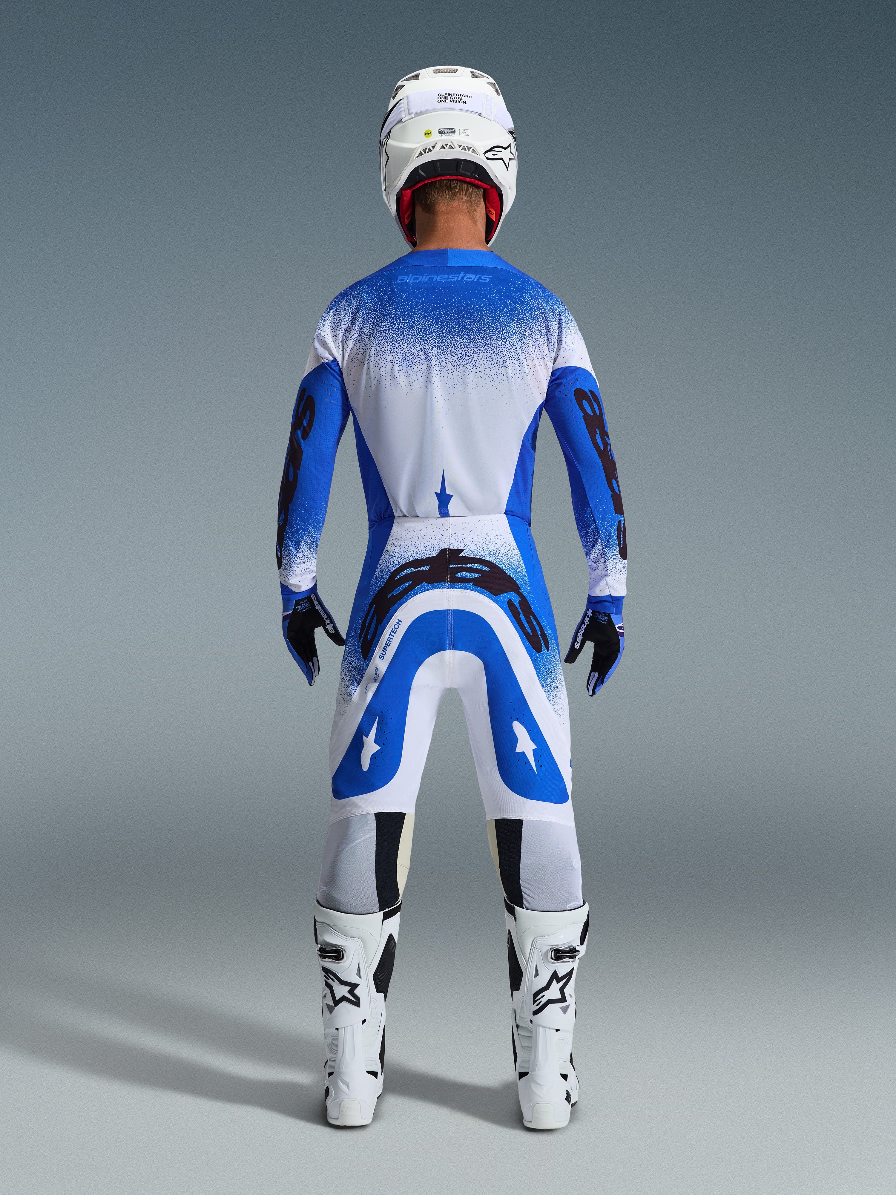 Motocross-Fahrer von hinten in kompletter Rennausrüstung, Alpinestars 2026 Supertech Scenz Hose in Ucla-Blau und Weiß, passendes blau-weißes Trikot mit gesprenkeltem Muster, weißer Helm, weiß-schwarze Stiefel, abgestimmtes Outfit für den professionellen Rennsport
