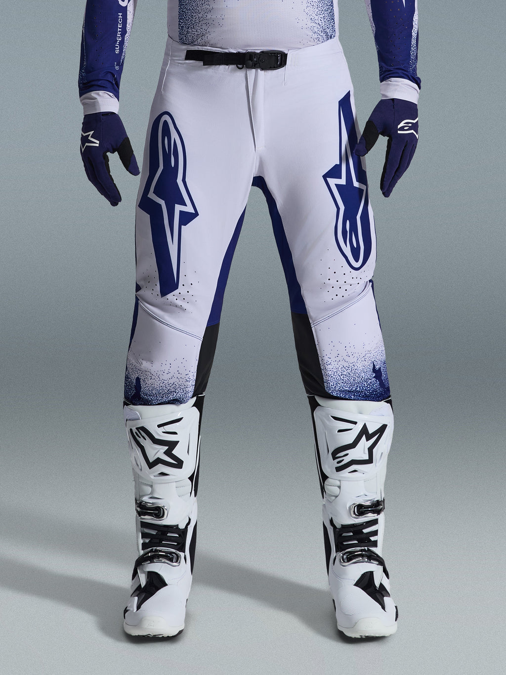 Alpinestars 2026 Supertech Scenz Hose, Motocross-Hose, Hellgrau und Dunkelblau, getragen mit weiß-schwarzen Motocross-Stiefeln und dunkelblauen Handschuhen, mit großen blauen Alpinestars-Logos auf den Oberschenkeln, SX1-Magnetverschluss und athletischer Passform für den Offroad-Rennsport