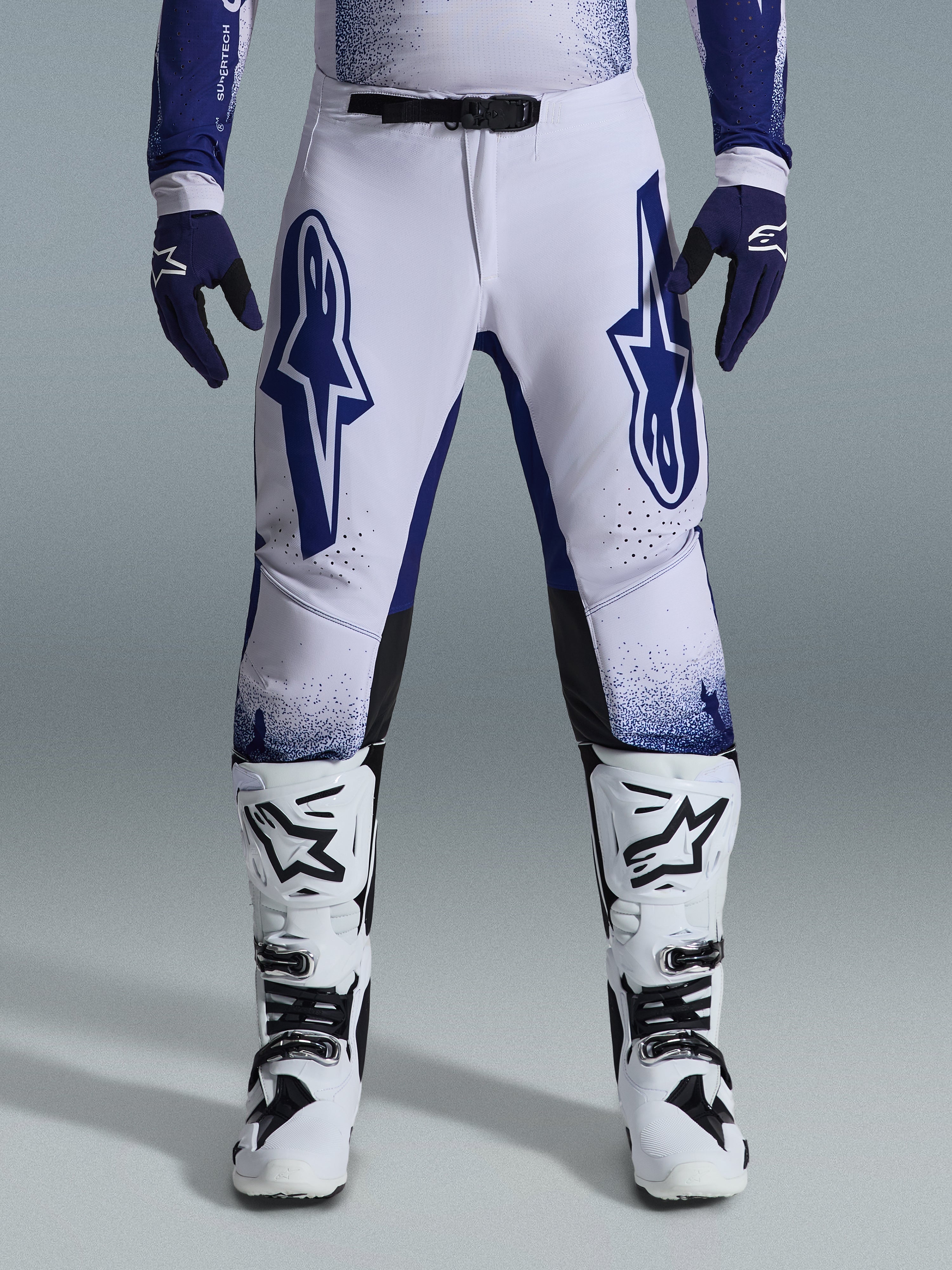 Alpinestars 2026 Supertech Scenz Hose, Motocross-Hose, Hellgrau und Dunkelblau, getragen mit weiß-schwarzen Motocross-Stiefeln und dunkelblauen Handschuhen, mit großen blauen Alpinestars-Logos auf den Oberschenkeln, SX1-Magnetverschluss und athletischer Passform für den Offroad-Rennsport
