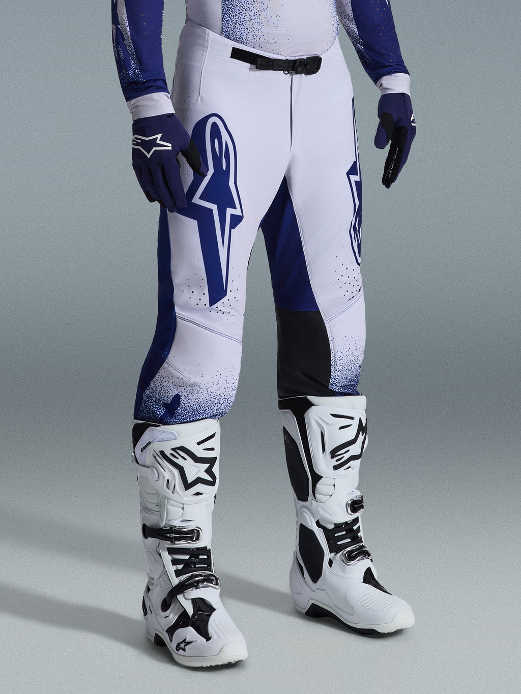 Alpinestars 2026 Supertech Scenz Hose, Motocross-Hose, Hellgrau und Dunkelblau, getragen mit weißen Motocross-Stiefeln und blauen Handschuhen, mit großen blauen Alpinestars-Logos auf den Oberschenkeln, schwarzen Knie-Panels und einem punktierten Grafikdesign, professionelle Offroad-Rennausrüstung