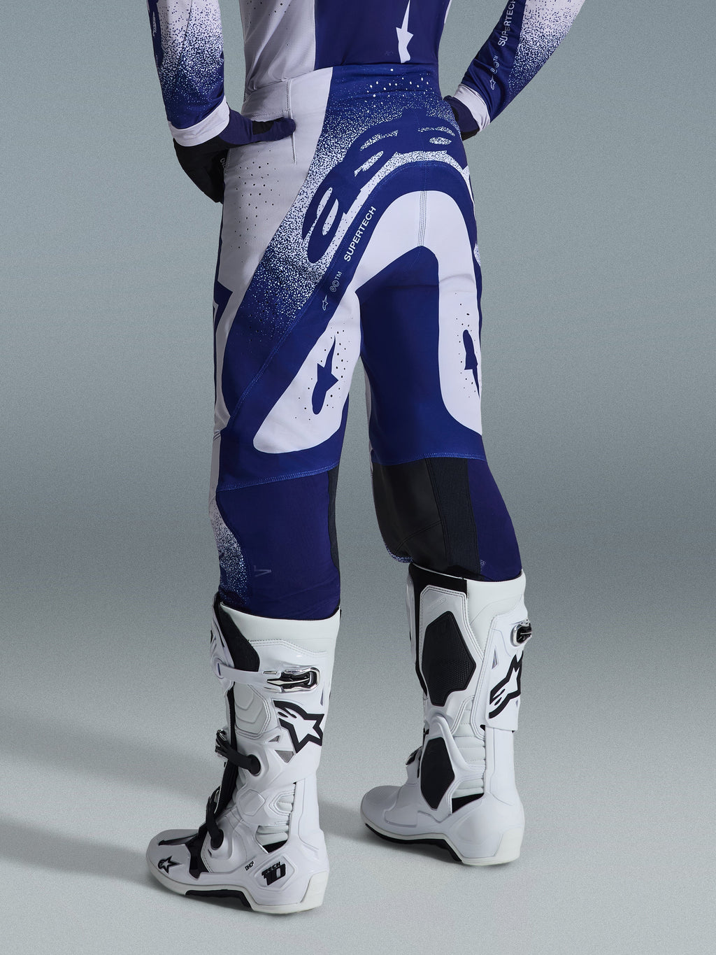Alpinestars 2026 Supertech Scenz Hose, Motocross-Hose, Hellgrau und Dunkelblau, Rückansicht getragen mit passendem Trikot und weißen Stiefeln, markante blaue Grafiken mit gesprenkelten Akzenten, athletische Performance-Passform für den Offroad-Rennsport.