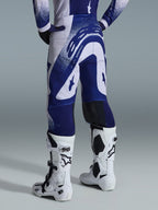 Alpinestars 2026 Supertech Scenz Hose, Motocross-Hose, Hellgrau und Dunkelblau, Rückansicht getragen mit passendem Trikot und weißen Stiefeln, markante blaue Grafiken mit gesprenkelten Akzenten, athletische Performance-Passform für den Offroad-Rennsport.