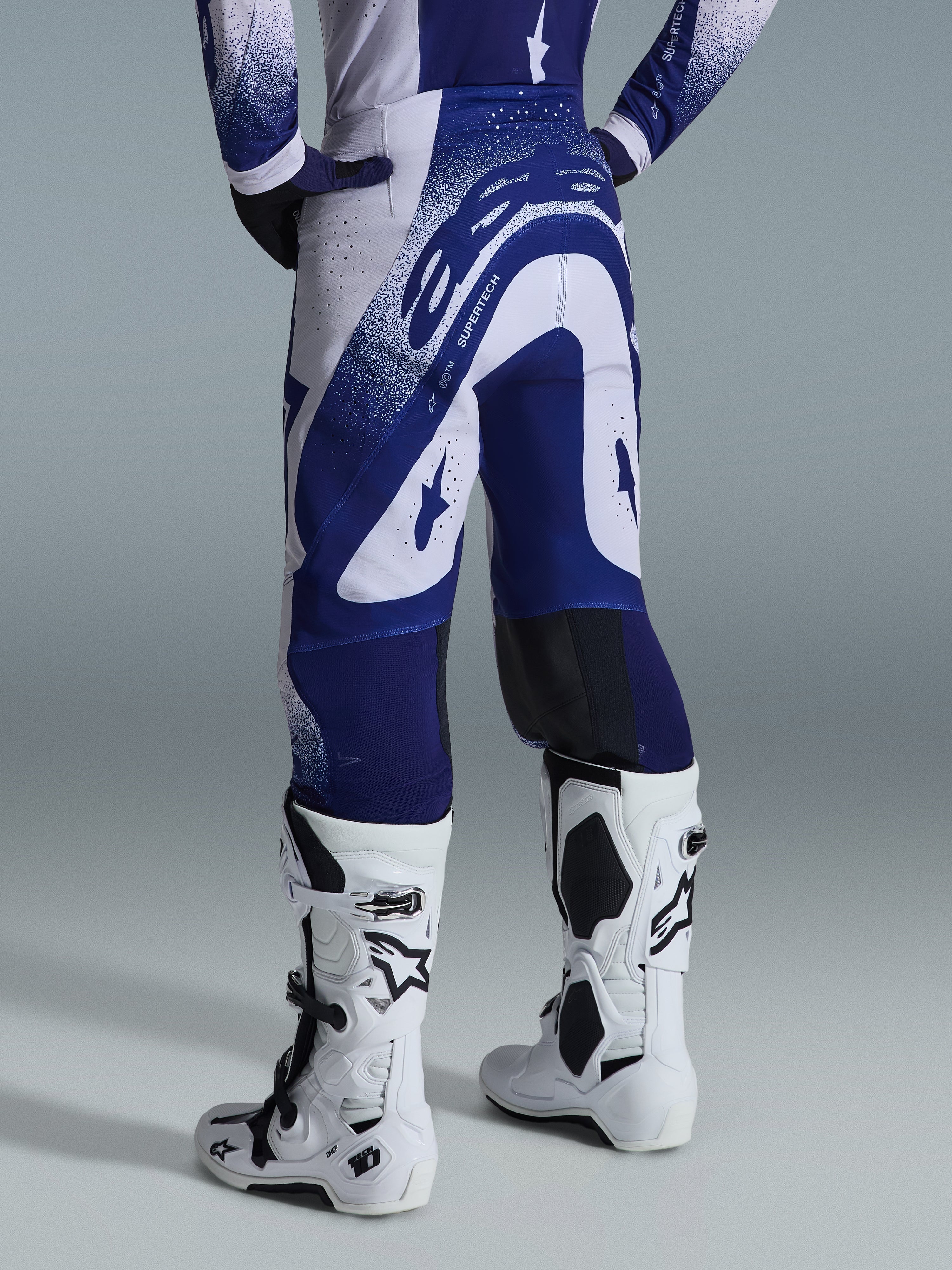 Alpinestars 2026 Supertech Scenz Hose, Motocross-Hose, Hellgrau und Dunkelblau, Rückansicht getragen mit passendem Trikot und weißen Stiefeln, markante blaue Grafiken mit gesprenkelten Akzenten, athletische Performance-Passform für den Offroad-Rennsport.