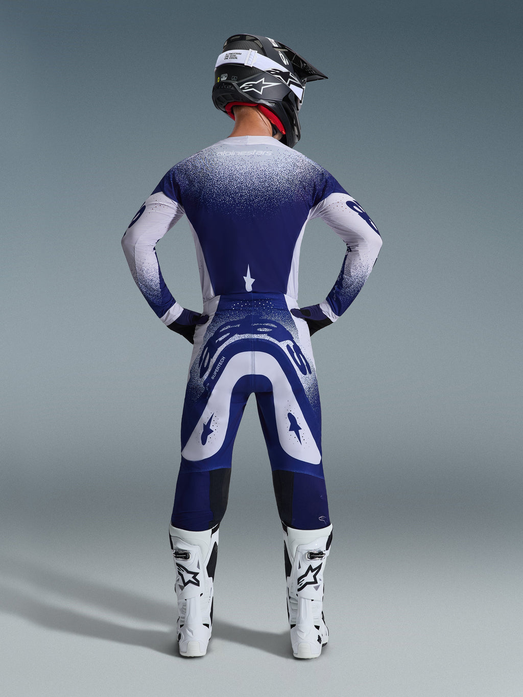 Alpinestars 2026 Supertech Scenz Hose, Motocross-Hose, Hellgrau und Dunkelblau, Rückansicht eines Fahrers in passender Ausrüstung mit gesprenkelten Farbverlaufsgrafiken, weiße Stiefel und schwarzer Helm, professionelle sportliche Passform für Offroad-Rennen
