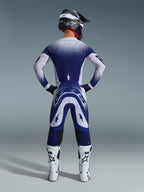 Alpinestars 2026 Supertech Scenz Hose, Motocross-Hose, Hellgrau und Dunkelblau, Rückansicht eines Fahrers in passender Ausrüstung mit gesprenkelten Farbverlaufsgrafiken, weiße Stiefel und schwarzer Helm, professionelle sportliche Passform für Offroad-Rennen