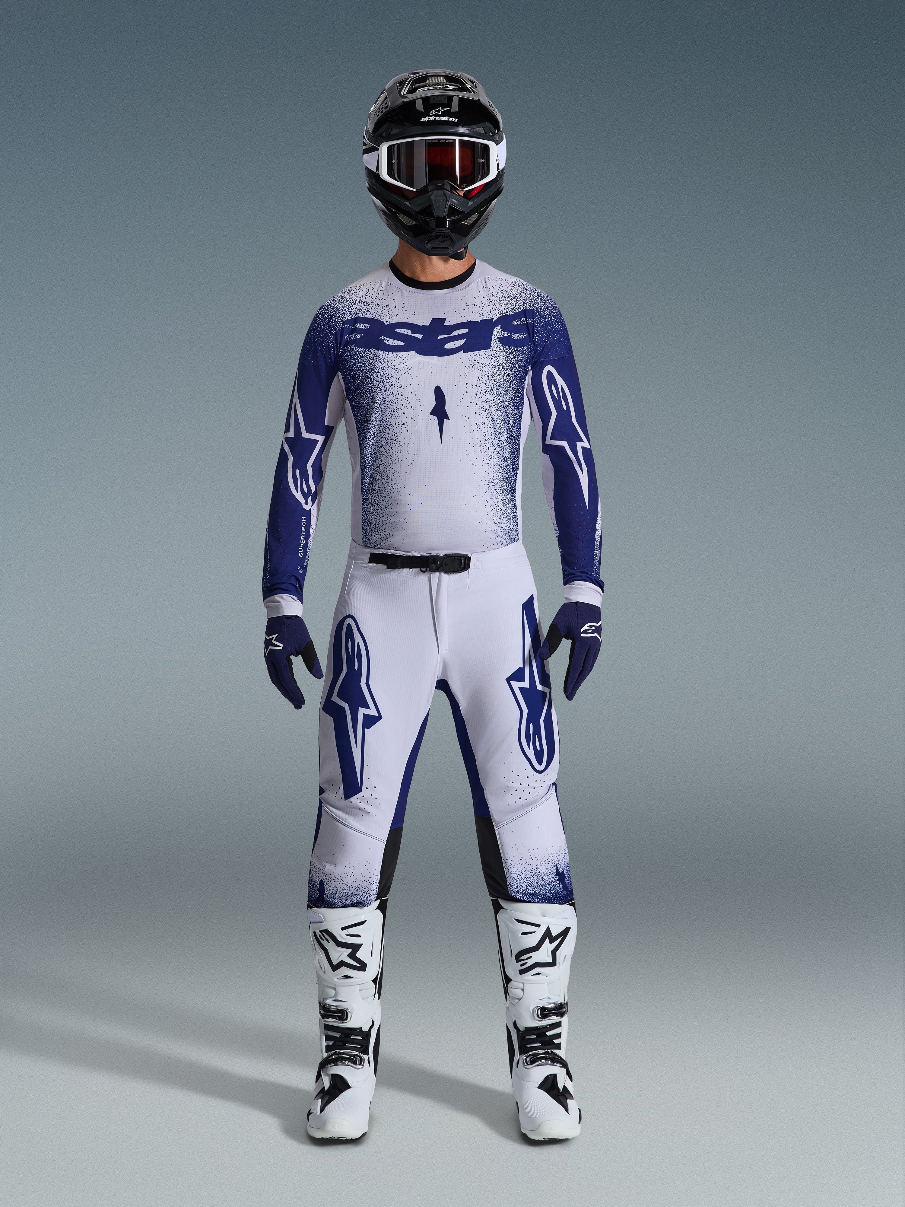 Motocross-Fahrer im Alpinestars 2026 Supertech Scenz Gear Set, Hellgrau und Dunkelblau, inklusive Trikot und Hose mit passenden blauen Handschuhen, weiß-schwarzen Stiefeln und einem schwarzen Helm mit weißer Brille, koordiniertes Outfit für professionellen Offroad-Rennsport