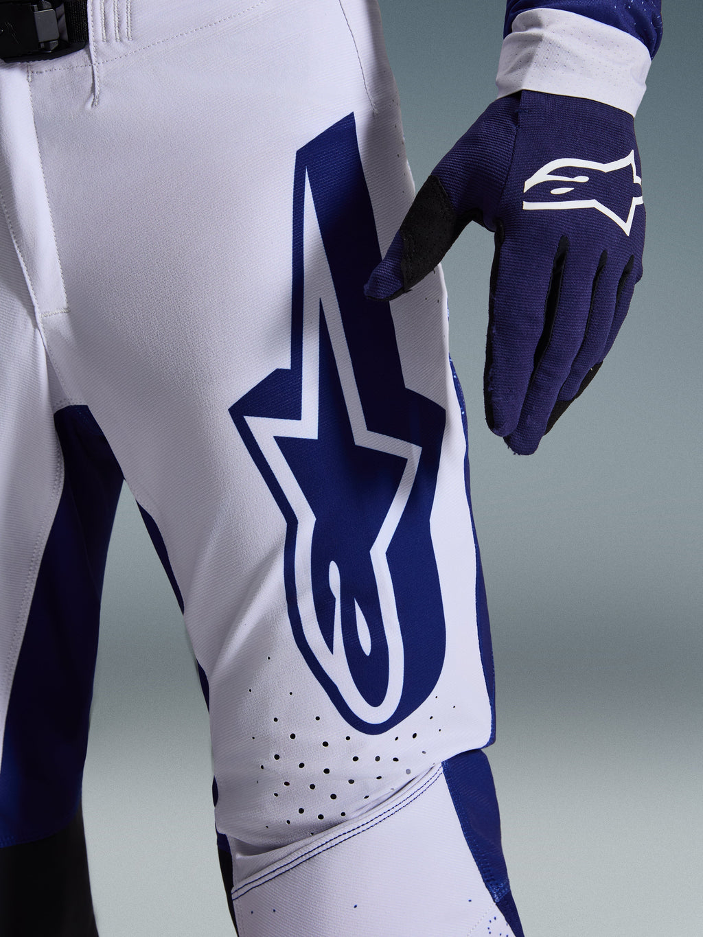 Alpinestars 2026 Supertech Scenz Hose, Motocross-Hose, Hellgrau und Dunkelblau, Nahaufnahme des Oberschenkels mit großem dunkelblauem Alpinestars-Logo, perforierte Belüftungszonen und Stretch-Ripstop-Gewebe, getragen mit passendem dunkelblauem Motocross-Handschuh