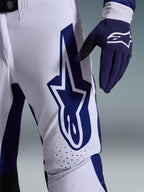 Alpinestars 2026 Supertech Scenz Hose, Motocross-Hose, Hellgrau und Dunkelblau, Nahaufnahme des Oberschenkels mit großem dunkelblauem Alpinestars-Logo, perforierte Belüftungszonen und Stretch-Ripstop-Gewebe, getragen mit passendem dunkelblauem Motocross-Handschuh