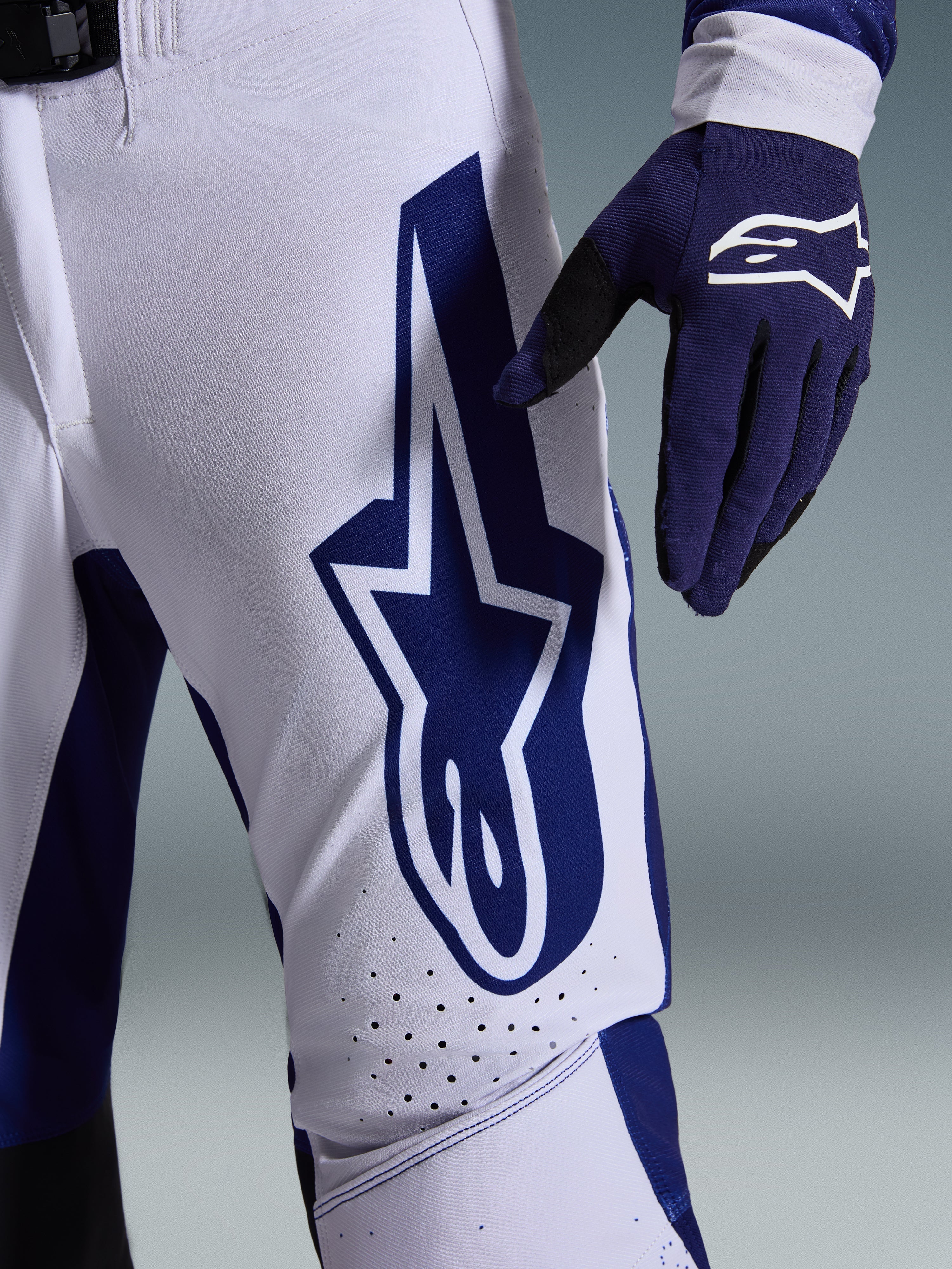 Alpinestars 2026 Supertech Scenz Hose, Motocross-Hose, Hellgrau und Dunkelblau, Nahaufnahme des Oberschenkels mit großem dunkelblauem Alpinestars-Logo, perforierte Belüftungszonen und Stretch-Ripstop-Gewebe, getragen mit passendem dunkelblauem Motocross-Handschuh
