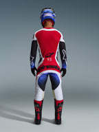 Alpinestars 2026 Techstar Knif Hose, Motocross-Hose, Schwarz, Weiß, Rot und Blau, Rückansicht eines Fahrers in kompletter Ausrüstung mit passendem Trikot, blauem Helm und schwarz-roten Stiefeln, mit markantem Alpinestars-Branding auf der Gesäßpartie.