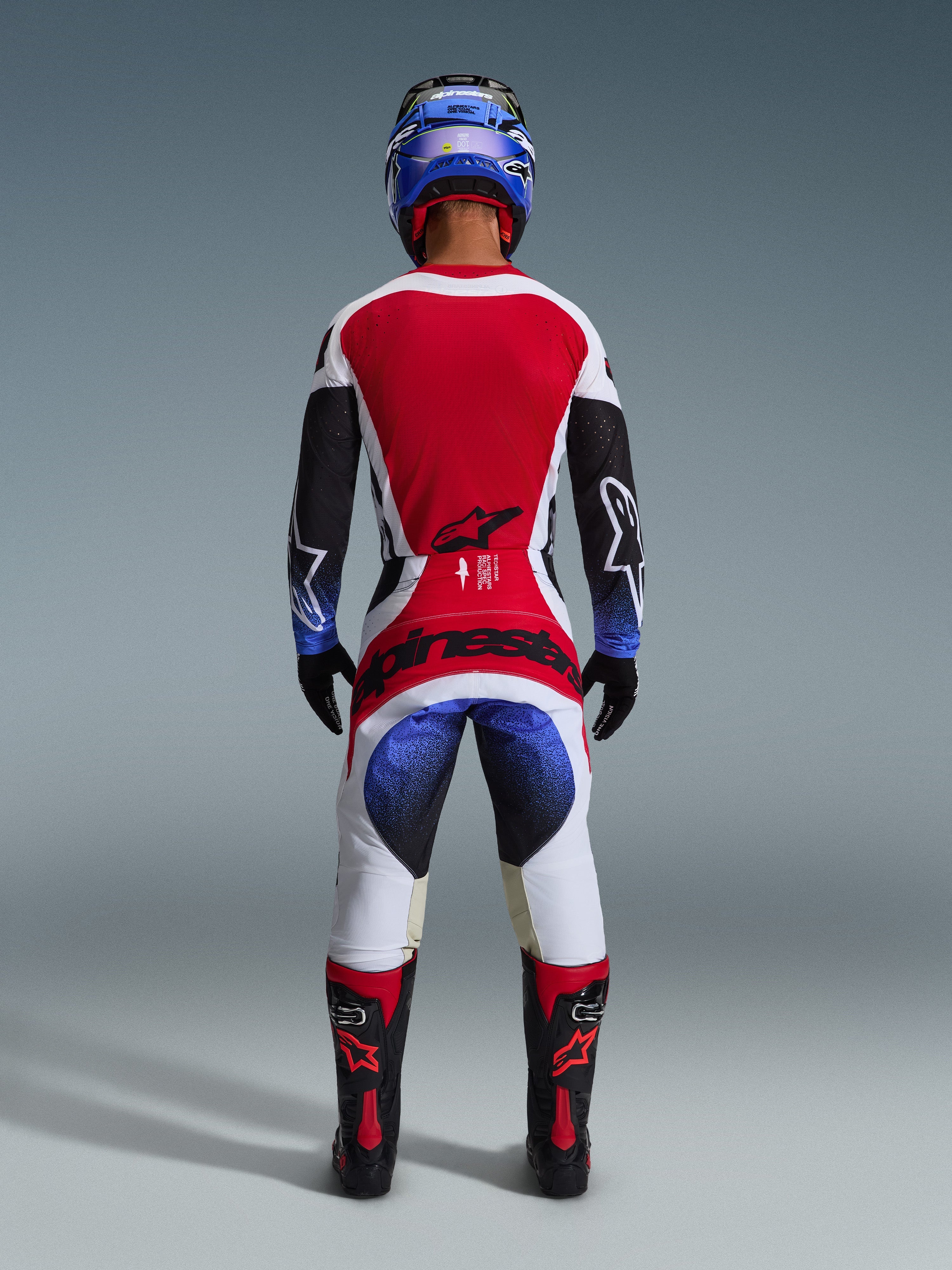 Alpinestars 2026 Techstar Knif Hose, Motocross-Hose, Schwarz, Weiß, Rot und Blau, Rückansicht eines Fahrers in kompletter Ausrüstung mit passendem Trikot, blauem Helm und schwarz-roten Stiefeln, mit markantem Alpinestars-Branding auf der Gesäßpartie.