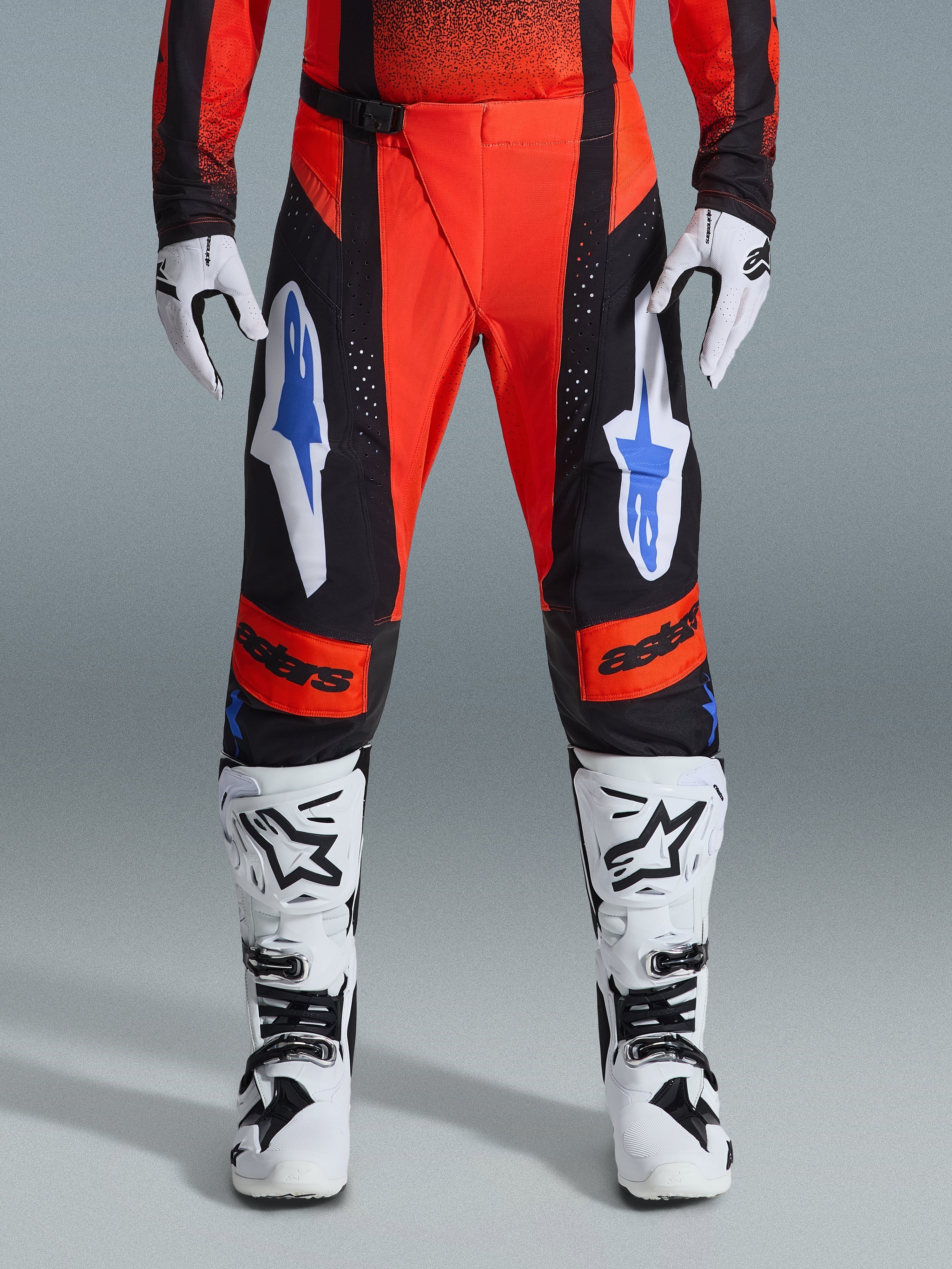 Alpinestars 2026 Techstar Knif Hose, Motocross-Hose, Orange, Schwarz und UCLA Blau, getragen mit passendem Jersey, weiß-schwarzen Motocross-Stiefeln und Handschuhen, mit auffälligem Branding an den Oberschenkeln und Knien, athletische Rennpassform für Offroad-Rennsport