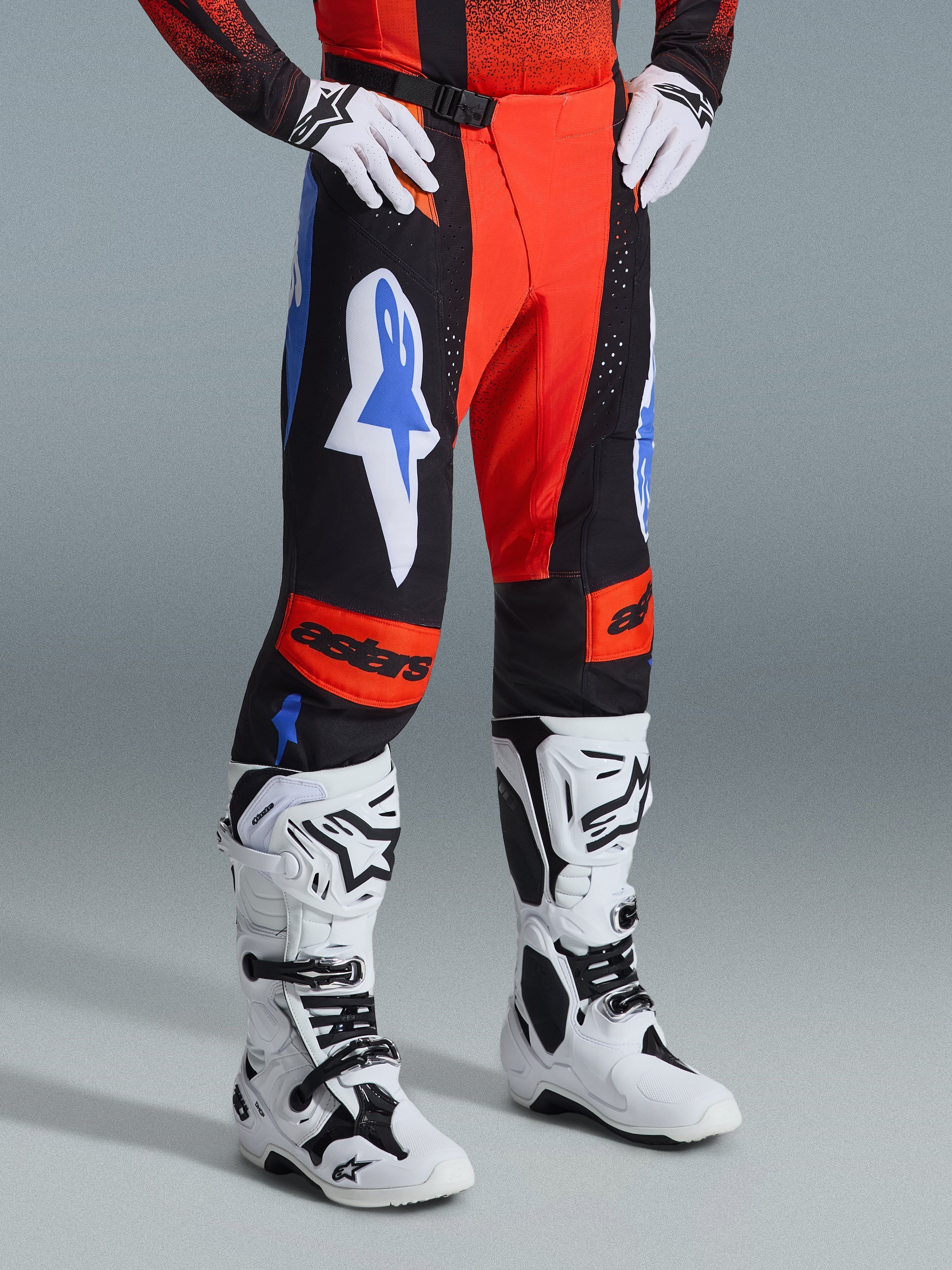 Alpinestars 2026 Techstar Knif Hose, Motocross-Hose, Orange, Schwarz und UCLA-Blau, getragen mit weißen Handschuhen und weiß-schwarzen Motocross-Stiefeln, mit markanten Logos auf den Oberschenkeln und orangen Kniepanels mit Astars-Branding.