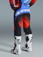 Alpinestars 2026 Techstar Knif Hose, Motocross-Hose, Orange, Schwarz und UCLA Blau, Rückansicht zeigt blaue Rückenpasse mit weißem Alpinestars-Logo, oranges Farbverlaufsmuster an den Beinen, kombiniert mit passendem Trikot und weißen Stiefeln für den Offroad-Rennsport