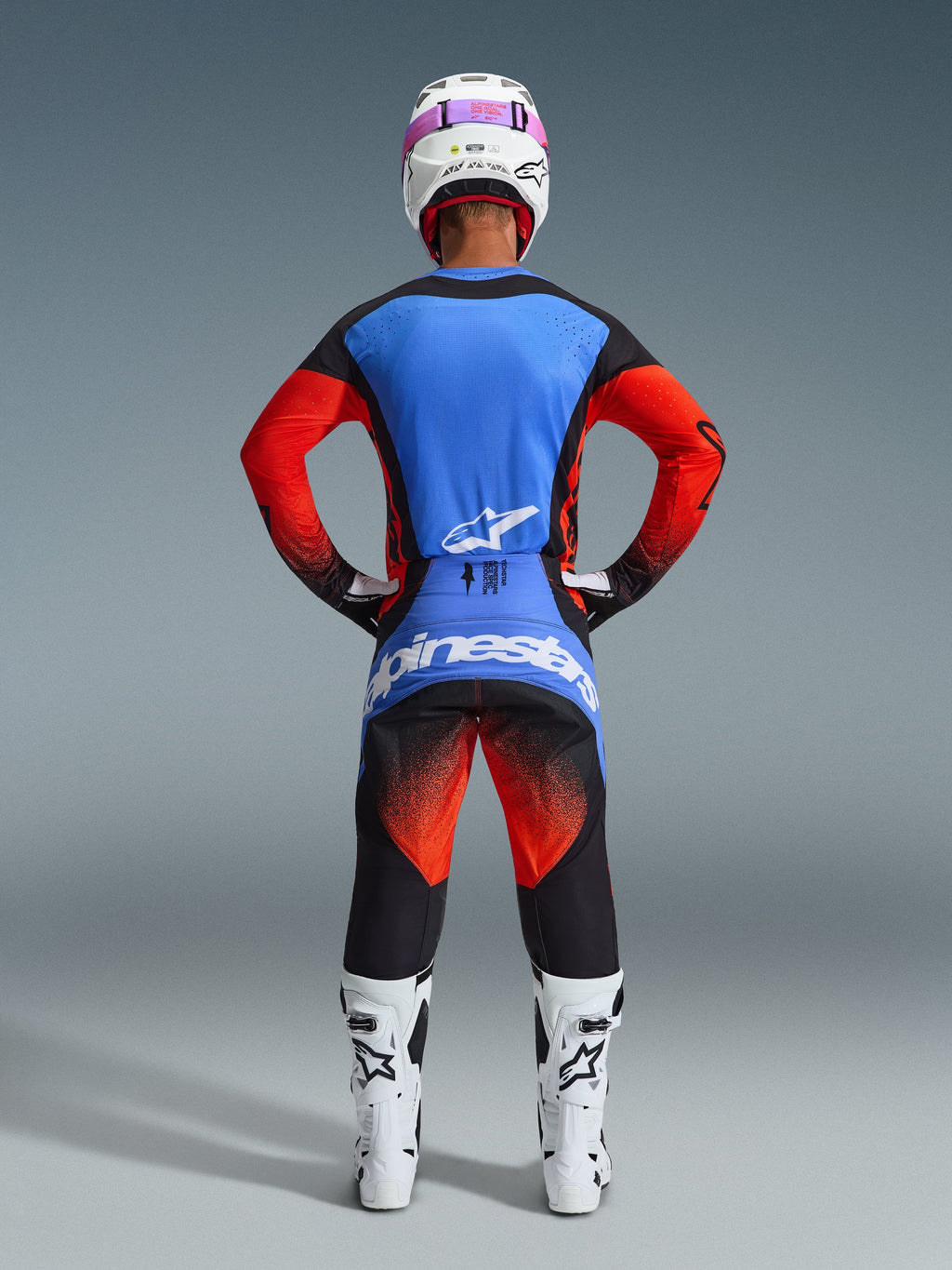 Motocross-Fahrer in der Rückansicht, trägt die 2026 Techstar Knif Hose in Orange, Schwarz und UCLA-Blau, kombiniert mit einem blau-orangen Trikot, weißem Helm mit pinker Brille und weißen Rennstiefeln, komplette Alpinestars Offroad-Wettkampfausrüstung