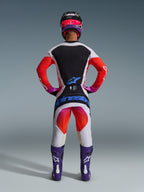 Motocross-Fahrer in kompletter 2026 Techstar Knif Rennausrüstung von hinten, hellgraues Trikot mit schwarzem Rückeneinsatz und orangefarbenen Ärmeln, passende weiß-orangefarbene Hose mit blauem Alpinestars-Branding, lila Stiefel und schwarz-pinker Helm, abgestimmtes professionelles Outfit für den Offroad-Rennsport
