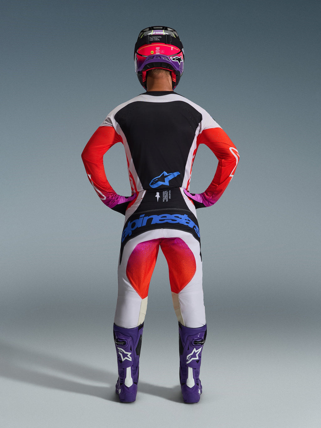Motocross-Fahrer in der Rückansicht mit 2026 Techstar Knif Hose und passendem Trikot, hellgraues und schwarzes Design mit leuchtend orangefarbenen und lila Akzenten, blaues Alpinestars-Logo auf der Gesäßpartie, lila Stiefel und Helm, professionelles Offroad-Rennausrüstungsset.