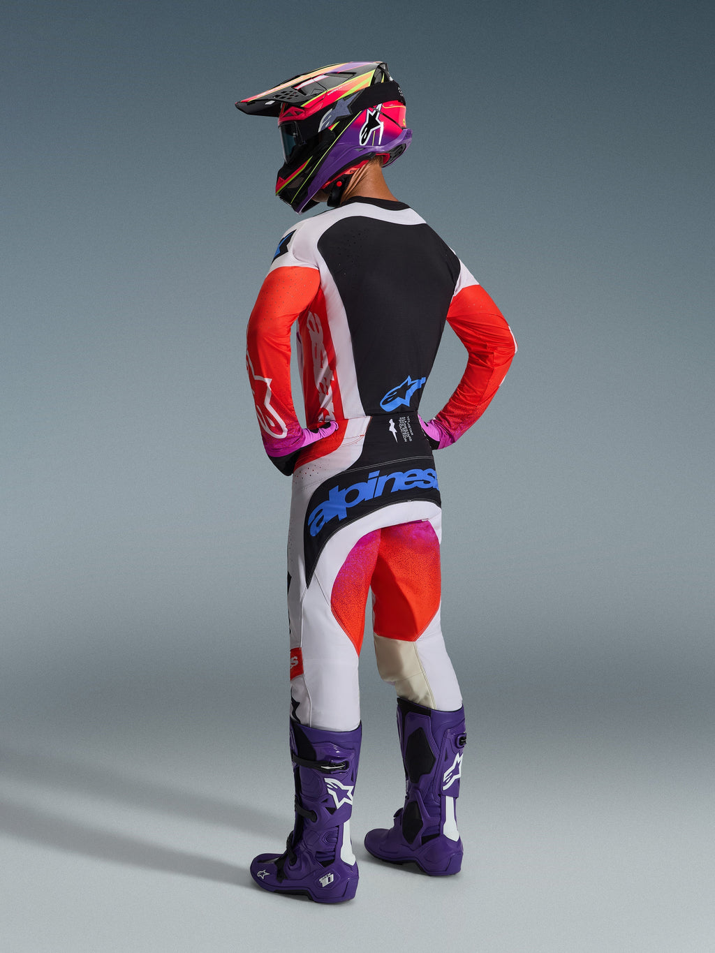 Motocross-Fahrer in der Rückansicht in kompletter Rennausrüstung, Alpinestars 2026 Techstar Knif Hose in Hellgrau und Orange Fluo mit blauem Branding, passendes Jersey mit orangefarbenen Ärmeln, lila Tech 10 Stiefel und ein farbenfroher Rennhelm, professionelles Offroad-Outfit