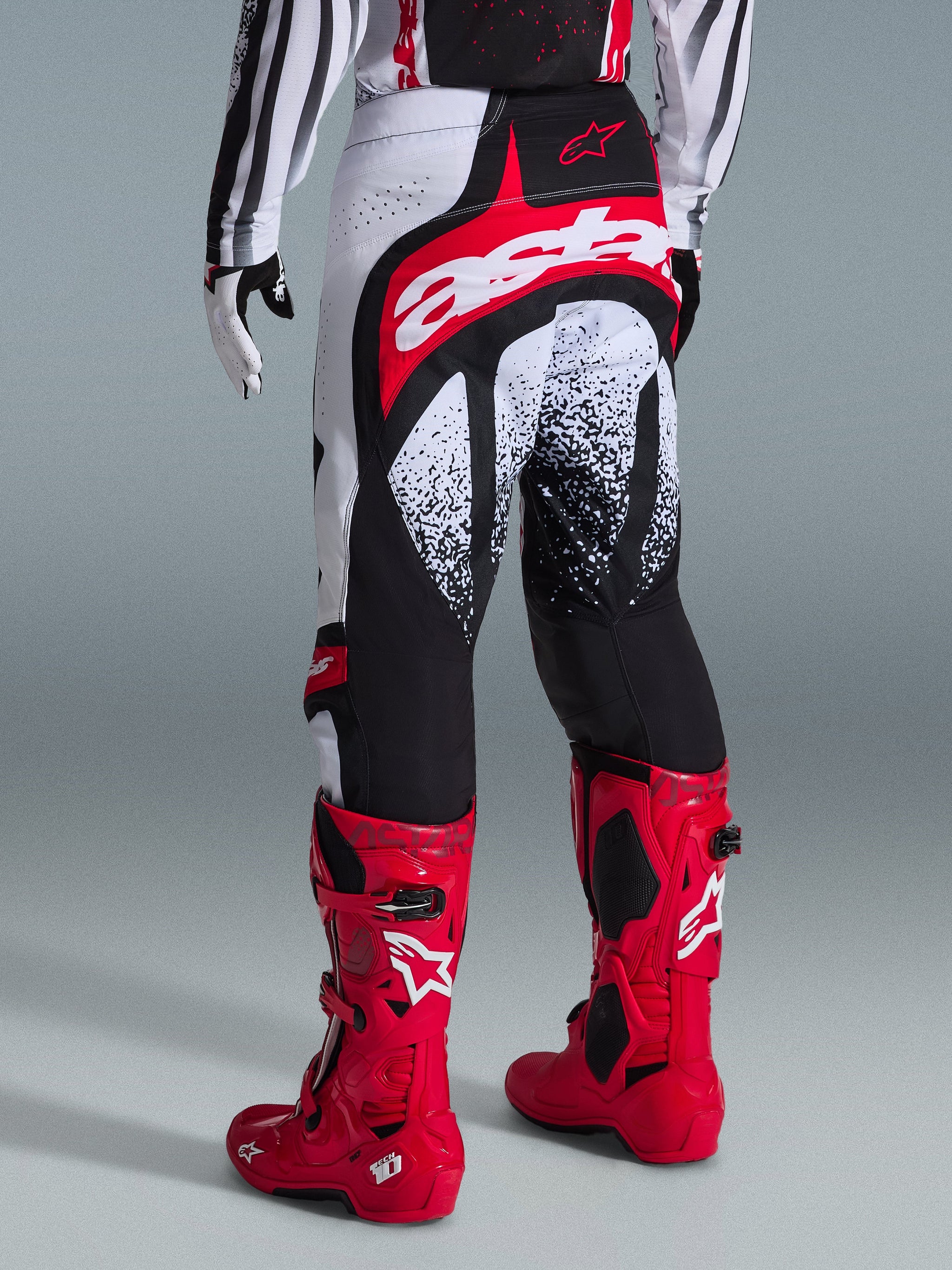 Alpinestars 2026 Techstar Nomur Pants, Motocross Pants, Black White and Red, rear view shown on a rider with matching red boots, featuring a bold red and white 'astars' logo on the seat and speckled graphic patterns, professional off-road racing gear